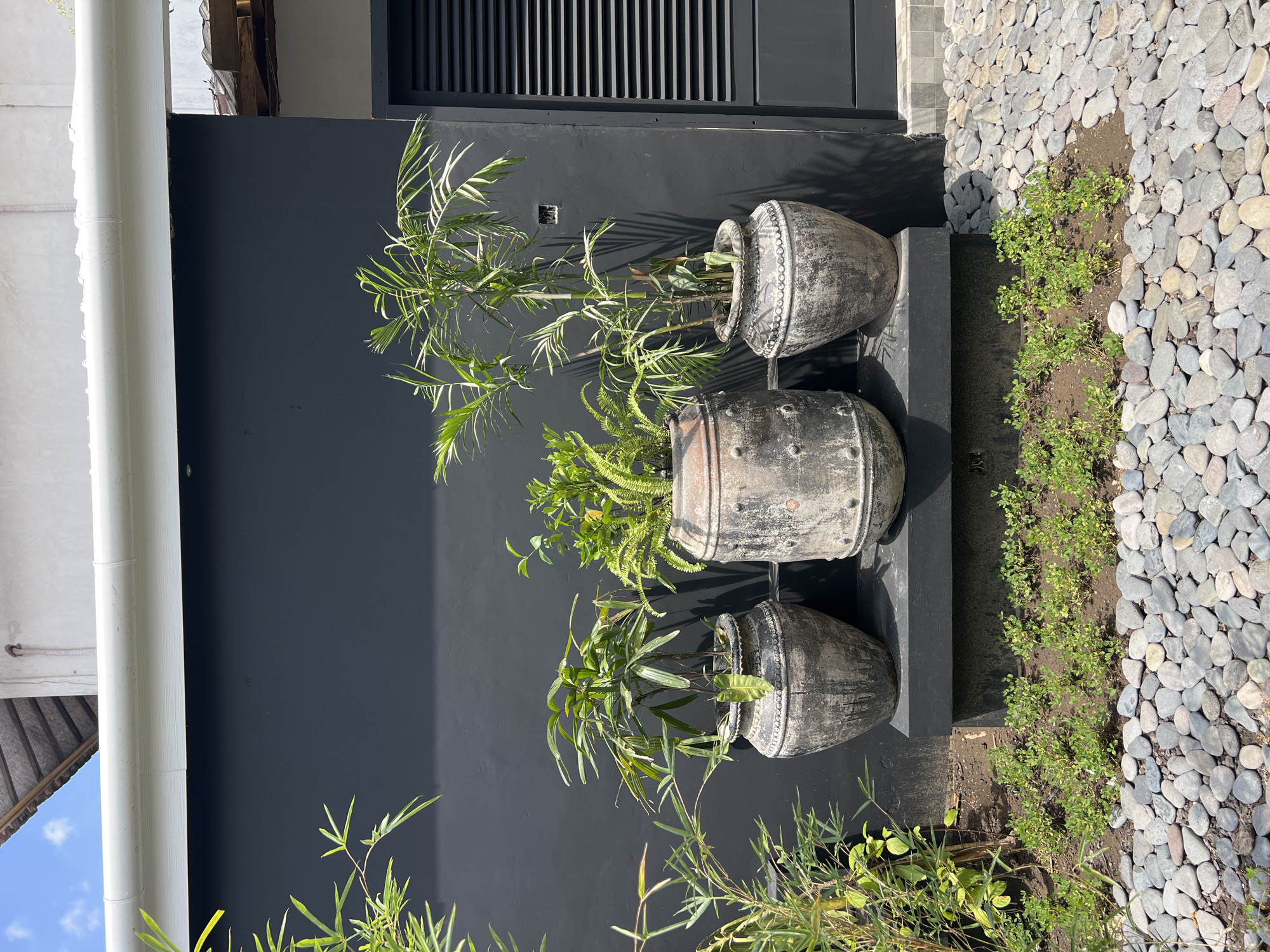 The image shows a vertical arrangement of three aged, gray, pot-like containers holding various plants, placed against a dark gray wall. The topmost container holds a plant with feathery leaves, the middle one a fern-like plant, and the bottom one another plant with slender leaves. To the right of the pots, there is a strip of soil containing small green plants, bordering a bed of smooth, gray stones. The lower part of the image features bamboo-like plants growing near the bottom pot. A white gutter is visible on the left edge of the wall. Above the pots, the dark gray wall features a louvered section. The location is North Kuta, Indonesia.