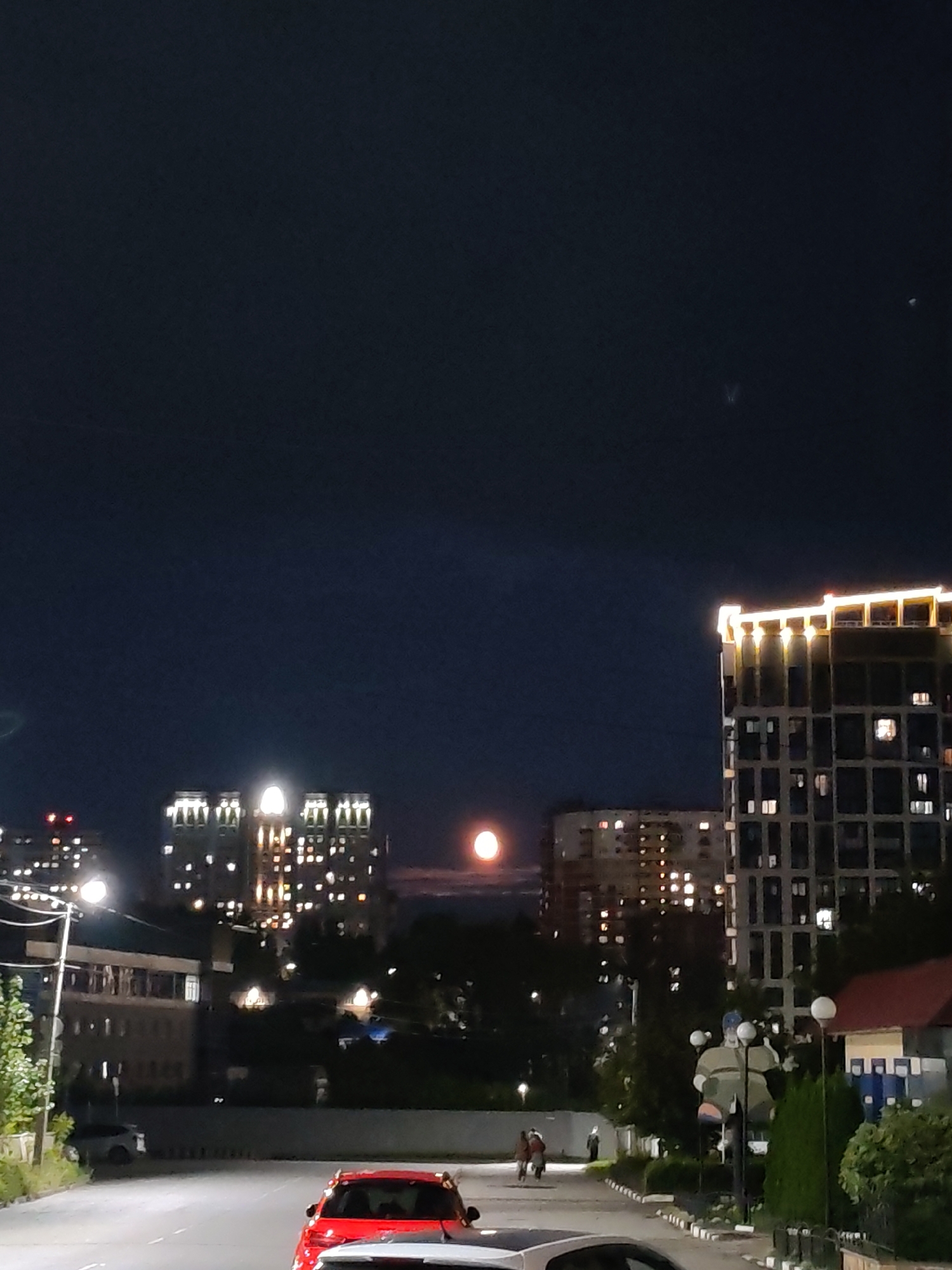 The image depicts a nighttime scene in Izhevsk, Russia, focusing on a street with several buildings and the moon visible in the sky.
In the foreground, a red car is parked on the right side of the street, partially visible. Further down the street, a white car is also present. The street itself appears to be paved, with white lines marking lanes. Streetlights are illuminated, casting light onto the road and surrounding areas.
Several buildings are visible in the mid-ground and background. To the right, a modern, tall building with a grid-like facade is illuminated by lights, with many windows showing interiors. Above this building, the sky is dark, with a bright, round moon positioned slightly left of center. To the left of this tall building, another, shorter building is visible, also with illuminated windows. Further to the left, a series of connected buildings with decorative, illuminated tops stand out.
In the mid-ground, trees and other greenery are present, contributing to the urban landscape. A few figures are seen walking on a sidewalk or pathway between the buildings. One prominent object is a large, stylized statue of a person in a green and beige outfit, with spheres on poles behind it.
The overall impression is an urban environment at night, with artificial lights from buildings and streetlights competing with the natural light of the moon. The scene appears calm and relatively quiet.