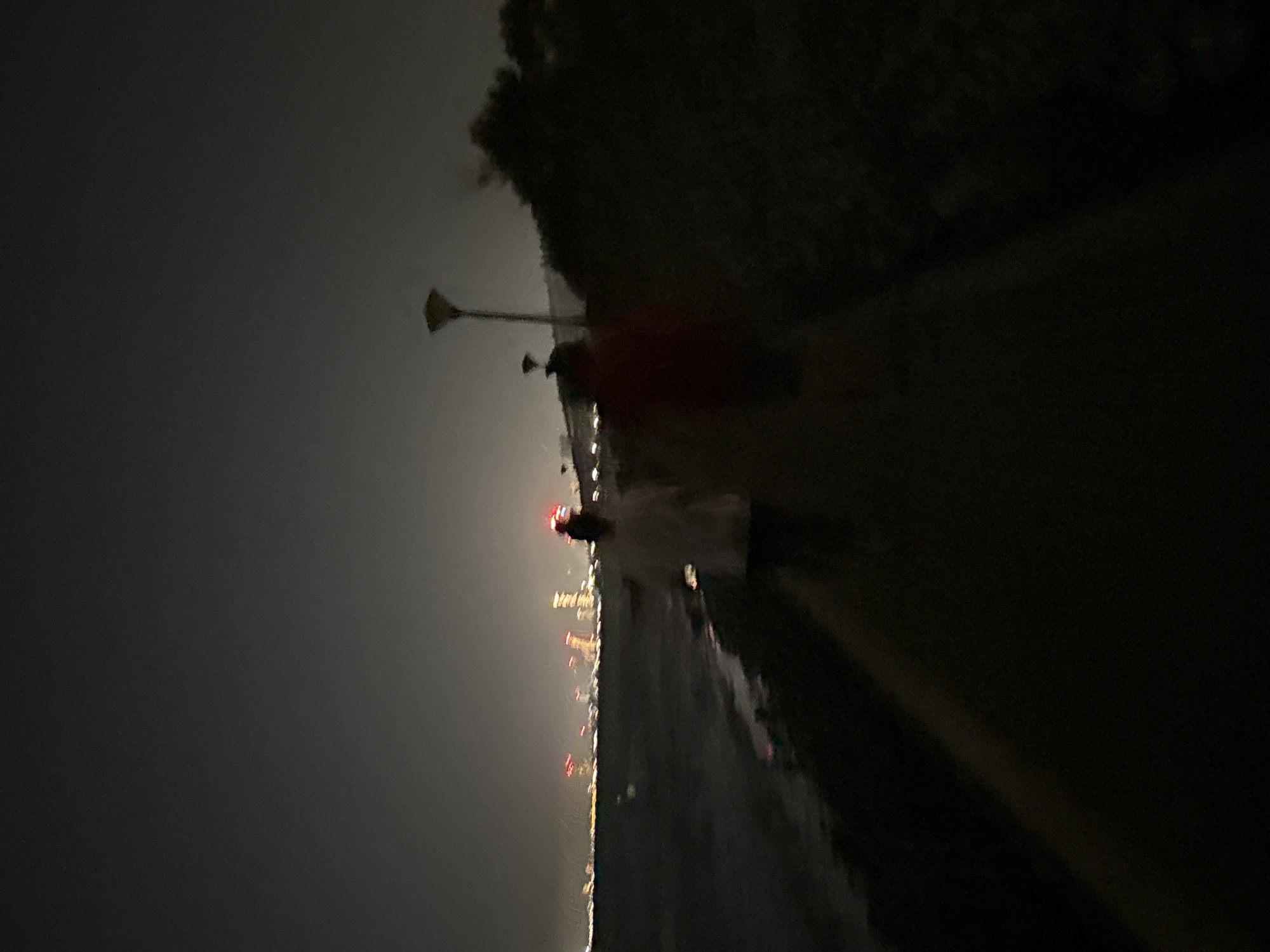 Two figures are visible on a paved path at night, with the sea on their left and dense foliage on their right. The figure on the left, wearing a white top, is slightly ahead of the figure on the right, who is wearing a red top. In the distance, a cityscape is illuminated by lights, with tall buildings and red and yellow lights visible. A lamppost with a distinctive shade is present on the right side of the path, near the foliage. The overall scene suggests a public walkway or promenade, potentially along the coast of Parekklisia Community, Cyprus, given the city lights in the background.