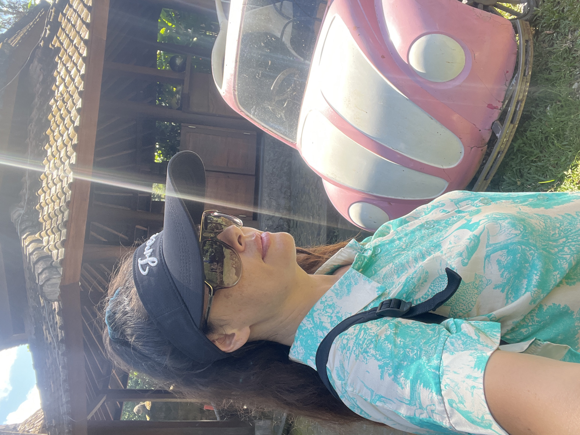 A medium shot captures a woman from the chest up, posed in profile, facing towards the right side of the frame. She is wearing a black visor with white lettering, oversized sunglasses, and a short-sleeved button-down shirt featuring a white base with a teal and white abstract pattern. A black backpack strap is visible across her shoulder. Her hair is dark and pulled back into a ponytail.
The woman is situated in front of a vintage pink Volkswagen Beetle with a cream-colored stripe down the hood. The car appears to be a decorative object, possibly for display, as it is positioned on what looks like a metal base.
In the background, a traditional Balinese-style building with a tiled roof and wooden pillars is partially visible. The environment is outdoors, with lush green grass and foliage in the foreground and background, suggesting a warm, sunny day. The lighting indicates a bright, possibly midday, sun with lens flare present in the upper left quadrant of the image. The overall scene suggests an outdoor attraction or tourist spot in Denpasar, Indonesia, likely a cultural or recreational area.