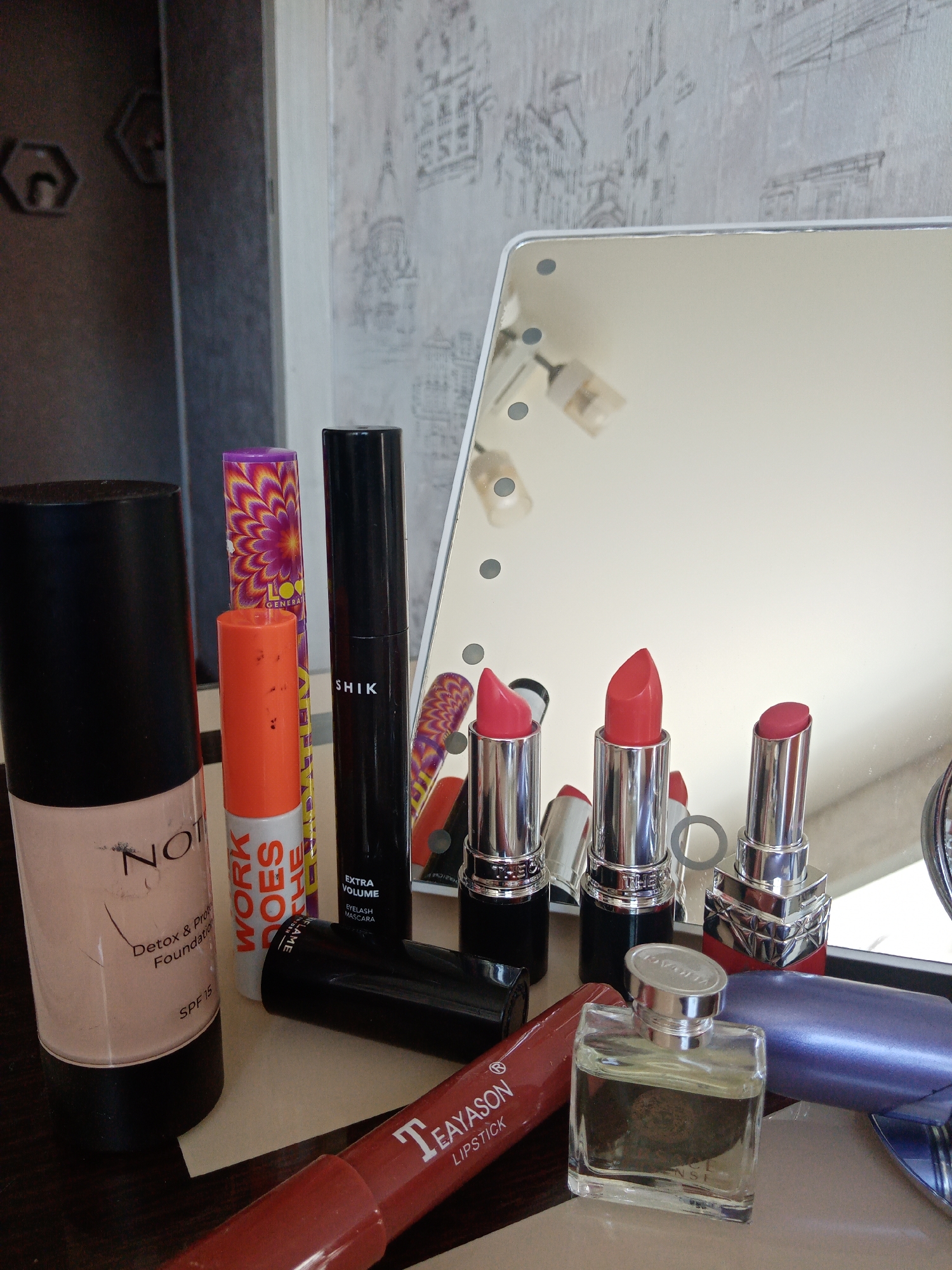 The image shows a collection of makeup items arranged on a surface, likely a vanity table. The items include: a bottle of "NOTE Detox & Protect Foundation SPF 15", an orange "WORK DOES THE FLAME" lip product, a purple "THE GENERAT" item with a firework design, a black "SHIK EXTRA VOLUME EYELASH MASCARA", several lipsticks in silver tubes with red and pink shades, a "TEAYASON LIPSTICK", and a small square bottle of "VERSACE" perfume. The background includes a mirror with built-in lights and wallpaper with a cityscape pattern. The location appears to be indoors, possibly in Elektrostal, Russia.