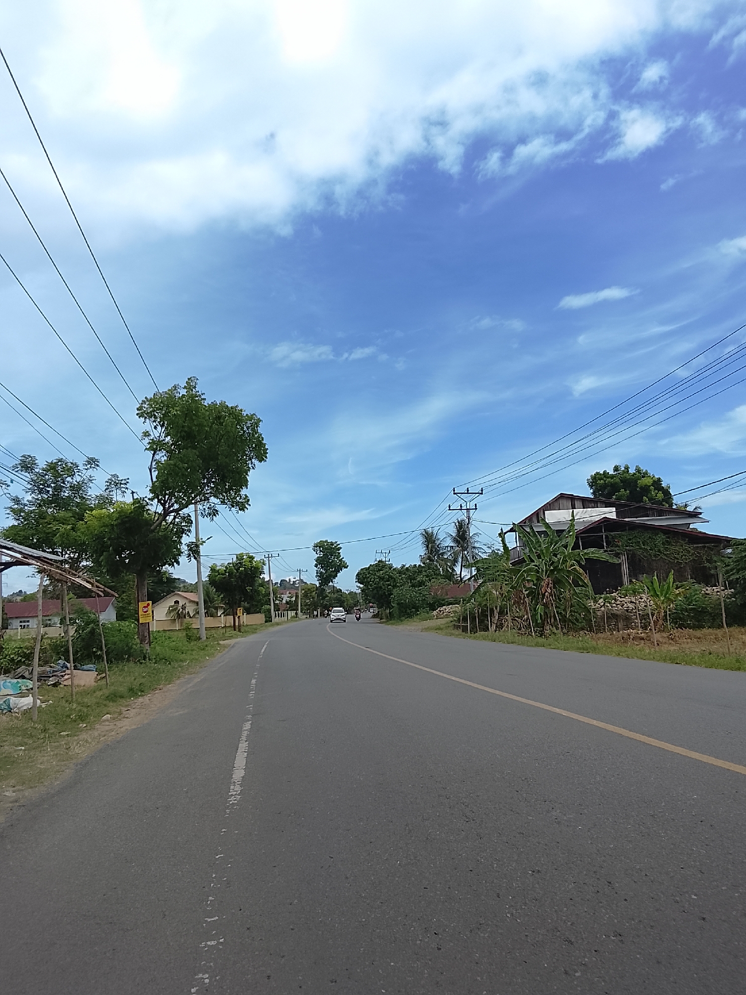 The image depicts a paved road in Bunot, Indonesia, under a bright blue sky with scattered white clouds. On the left side of the road, there are several trees and a small structure with a thatched roof. Further down the road, houses and more vegetation are visible. On the right side, there is another house with a covered porch, surrounded by trees and plants, and a stone wall in front. Utility poles with numerous wires stretch across the scene, following the path of the road. Two vehicles are present on the road in the distance, one white car and a motorbike. The road itself is asphalt with a broken white line marking the center and a solid yellow line on the right edge. The overall environment appears to be a rural or semi-rural area with residential buildings and natural elements.