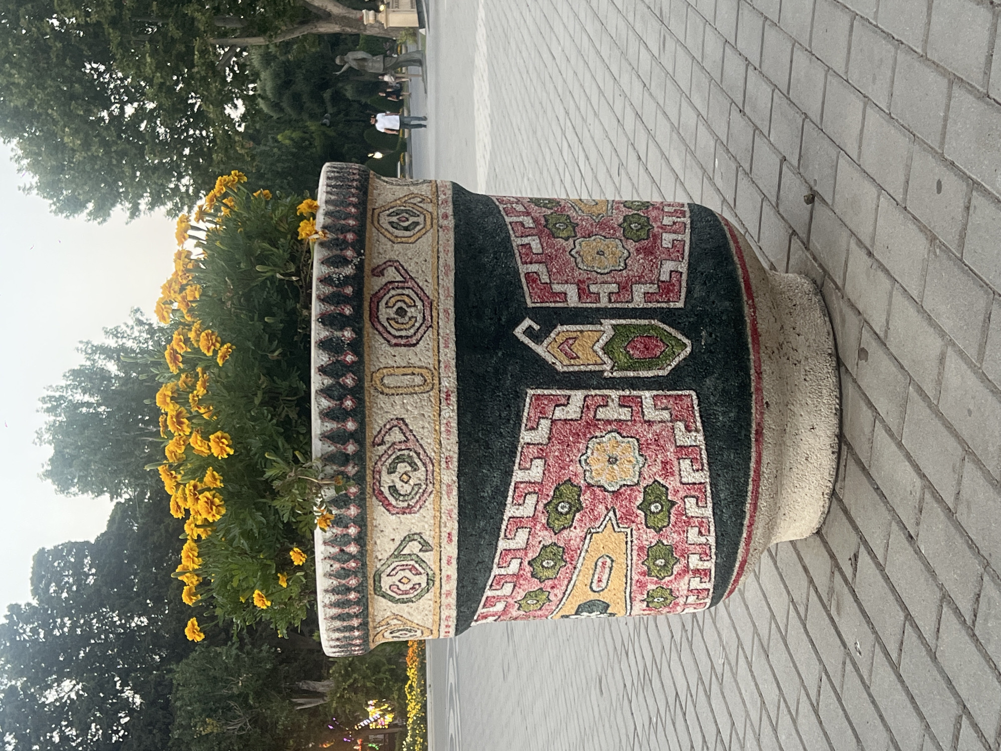 In the city of Sabail Raion, Azerbaijan, a large, ornate planter sits on a paved walkway. The planter is decorated with patterns resembling Azerbaijani carpets, featuring geometric shapes and floral motifs in red, green, yellow, and black. Bright yellow flowers bloom abundantly from the top of the planter. In the background, trees line a path, and a few people are visible walking. To the right, a statue of a man stands prominently. The sky is overcast, suggesting it might be late afternoon or early evening.