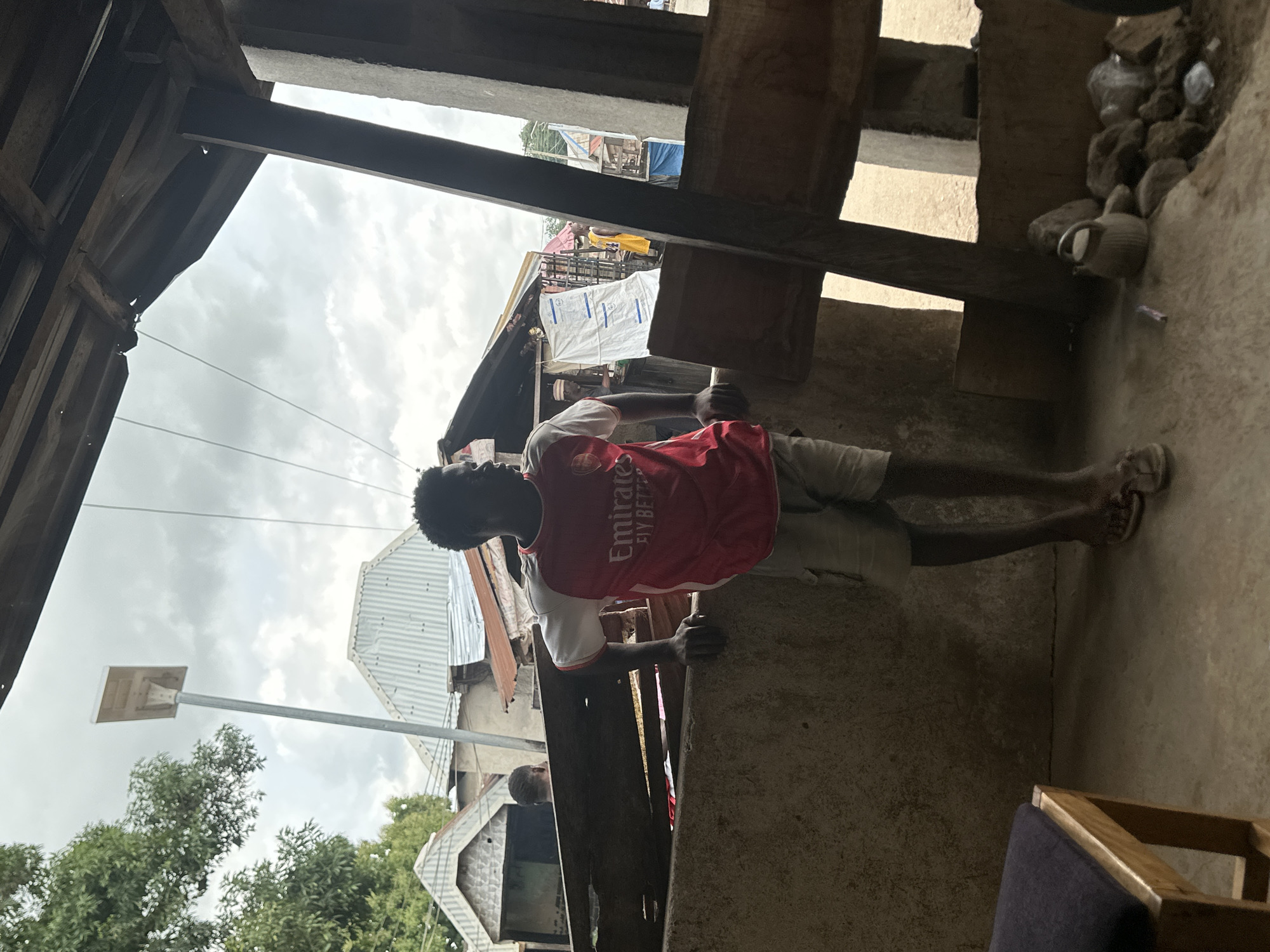 In Mararaba, Nigeria, a young Black male stands leaning on a concrete barrier, looking to his right. He wears a red and white short-sleeved t-shirt with "Emirates FLY BETTER" printed on the front, and light-colored shorts. His feet are bare save for simple flip-flops. The environment appears to be an outdoor market or residential area with corrugated metal roofs visible in the background. A solar-powered street light stands tall behind him. The foreground shows a concrete floor, part of a wooden stool with a dark cushion, and a collection of rocks and a small kettle. The overall scene is under an overcast sky.