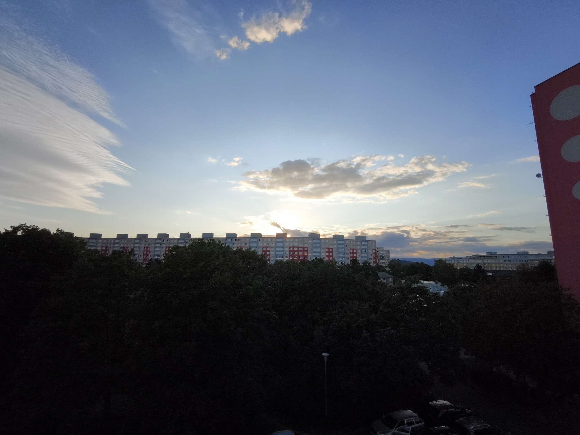 In the city of Most, Czechia, an urban landscape is captured during a sunset. The sky is a vivid blue, transitioning to lighter hues near the horizon, adorned with wispy, elongated clouds and scattered, darker clouds that catch the setting sun's light. A large, pink building with circular white patterns dominates the right side of the frame, partially obscuring a distant view of a long, multi-story residential building with a red and white facade, likely part of the Severní sídliště (Northern Housing Estate). In the foreground, dense, dark green foliage from trees creates a silhouetted barrier, with a few parked vehicles, primarily cars, visible in the lower right corner. The lighting suggests late afternoon or early evening, with the sun partially hidden behind clouds and casting a warm glow across the sky.