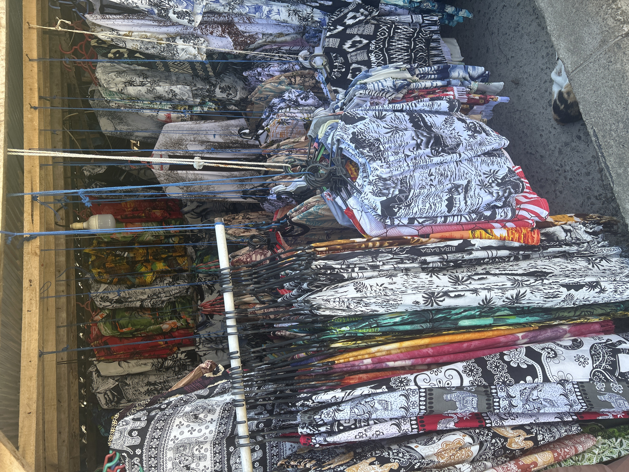 The image depicts a market stall in Bira, Indonesia, selling a variety of brightly colored and patterned clothing, predominantly short-sleeved shirts and possibly some dresses or skirts. The garments are hung from a wooden structure on multiple racks and suspended by blue cords. The patterns are varied, featuring tropical scenes, elephants, geometric designs, and abstract prints in black, white, red, green, yellow, and other vibrant hues. Some of the clothing appears to be made of light, breathable fabric, suggesting it is intended for a warm climate. In the lower right corner of the image, a calico cat is resting on a concrete surface, partially obscured by a low ledge. The ground beneath the stall is paved with concrete. The overall impression is of a bustling, colorful market setting characteristic of a tourist destination.