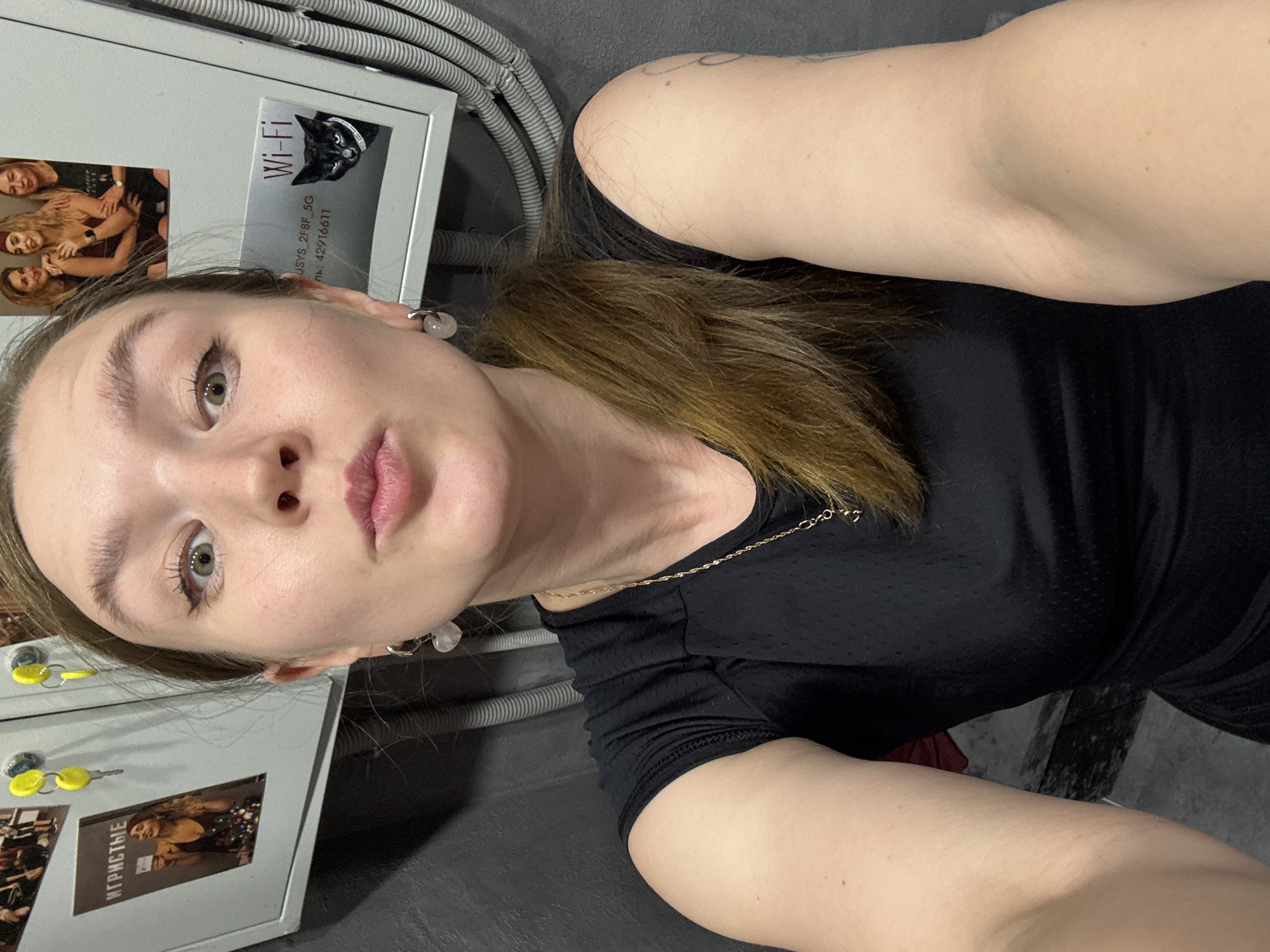 A young woman takes a close-up selfie in an indoor setting, likely in Izhevsk, Russia. The image is rotated, showing her face prominently on the left side, looking directly at the camera with a neutral expression. She has light-colored eyes, defined eyebrows, and light pink lips. Her long, brown hair is pulled back, and she wears light-colored stud earrings and a delicate gold-colored chain necklace over a black athletic-style top.
The background primarily consists of a light grey metal cabinet or utility box. Attached to this cabinet are several items: a "Wi-Fi" sticker featuring a cartoon black cat with a white collar, below which is visible text "JSYS_2F8F_5G" and "ль: 42916611". Also affixed are several small photographs, mostly featuring groups of women socializing. One partially visible framed image or magnet displays the Cyrillic word "ИГРИСТЫЕ" (meaning "Sparkling"). Nearby, two yellow keys or key covers hang on hooks. Above and to the right of the woman, grey corrugated conduits or cables run along a dark wall or ceiling. The overall setting appears to be a practical or utility area, possibly a back room or a technical space, rather than a decorative one.