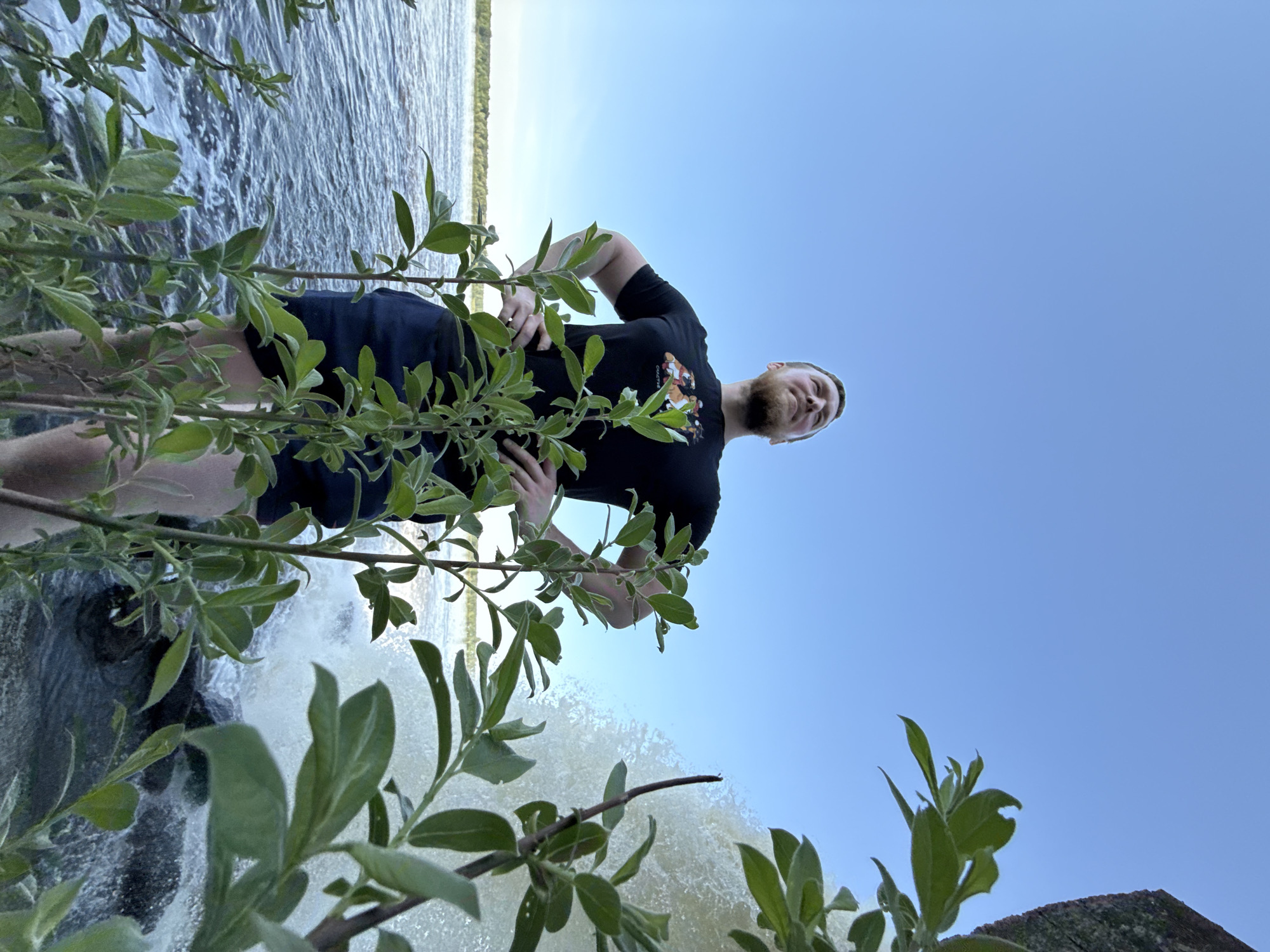 The image is oriented sideways, depicting a man standing near a waterfall. He is wearing a black t-shirt and dark shorts, with his hands on his hips. The background consists of a blue sky above and the flowing water of the waterfall behind him. Green foliage partially obscures the view in the foreground. The location is Novyi, Russia.