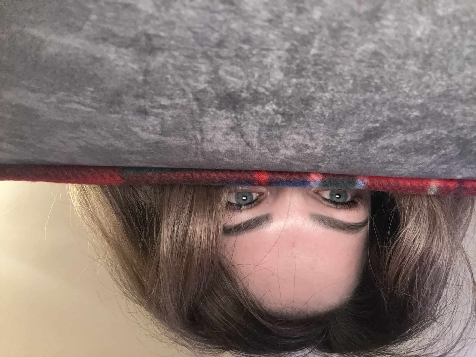 The image shows a woman's face from an upside-down perspective, peering from under a grey, possibly upholstered object trimmed with a red and blue plaid fabric. Her eyes are visible, along with part of her forehead, nose, and hair. The background beneath her head appears to be a light, indistinct surface. The location is unknown.