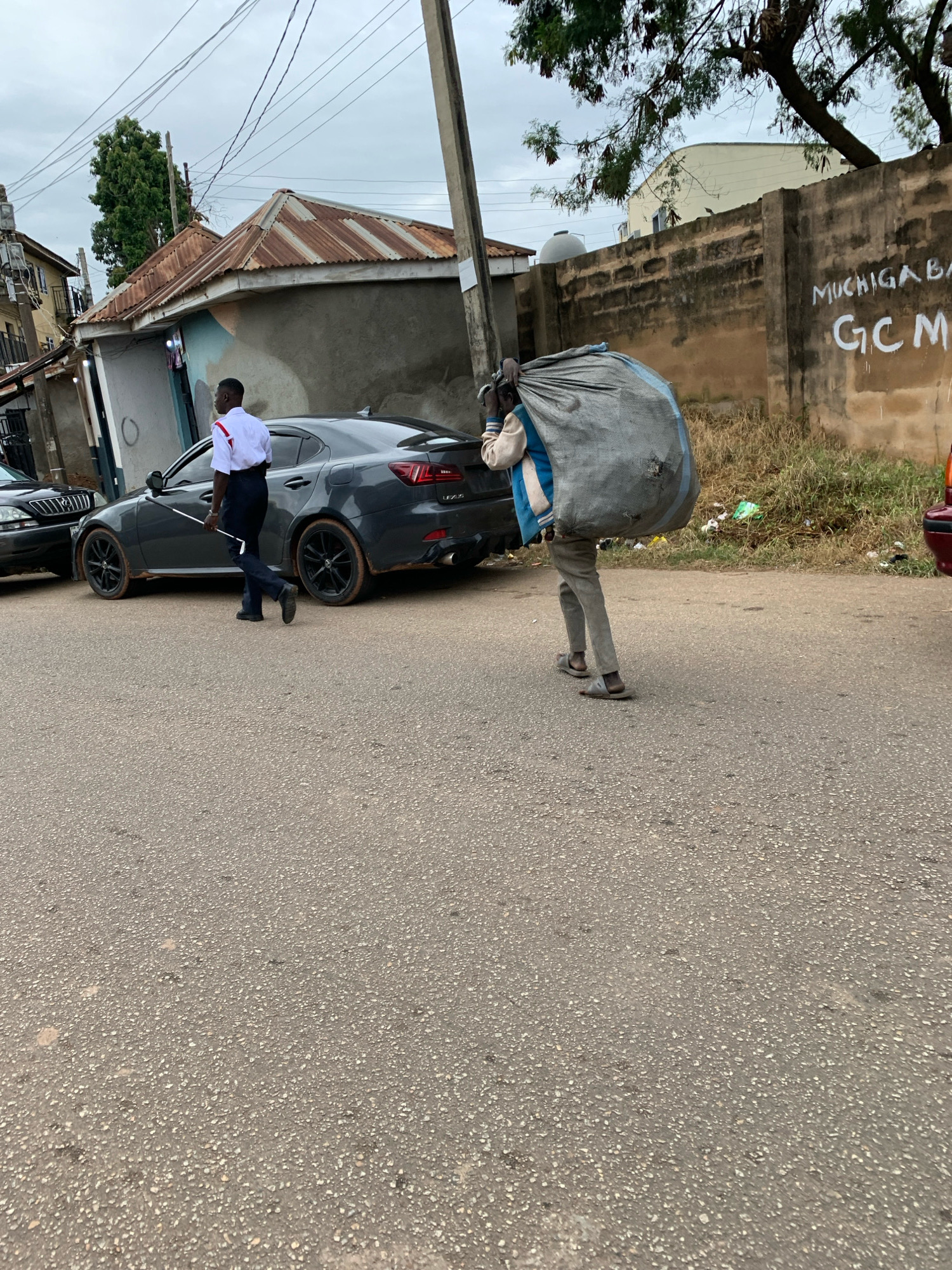 An overcast day in Jos, Nigeria, shows a street scene. On the left, a man in a white shirt with red trim and dark pants walks with a white stick. Beside him is a dark grey Lexus sedan with black rims, its tires dusty. To the right, a person is carrying a large, grey, lumpy bag on their back, walking towards the right of the frame, away from the camera. The person is wearing a light-colored jacket over a blue shirt and grey pants, with grey flip-flops. Behind them is a tall, roughly constructed brick wall with some graffiti. One message on the wall reads "MUCHIGABA" and below it "GCM". Adjacent to the wall is overgrown grass and scattered debris. In the background, the corrugated metal roof of a small building is visible, and beyond that, some trees and another building. A utility pole stands on the right side of the street.