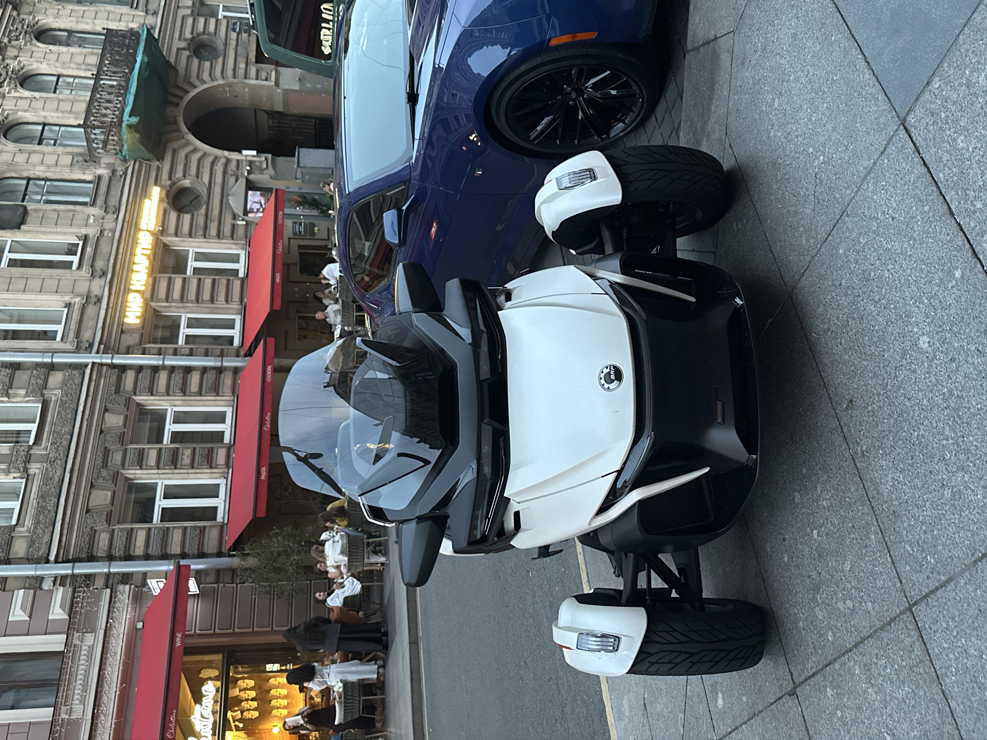 A white three-wheeled Can-Am Spyder is parked on a sidewalk in front of a building with red awnings and outdoor seating for a restaurant. To the right of the Spyder is a dark blue Chevrolet Camaro. The building appears to be old, with ornate architecture. Several people are seated at tables outside the restaurant, enjoying drinks and conversation. The street is paved with cobblestones. The overall scene depicts a vibrant urban setting in Saint Petersburg, Russia.