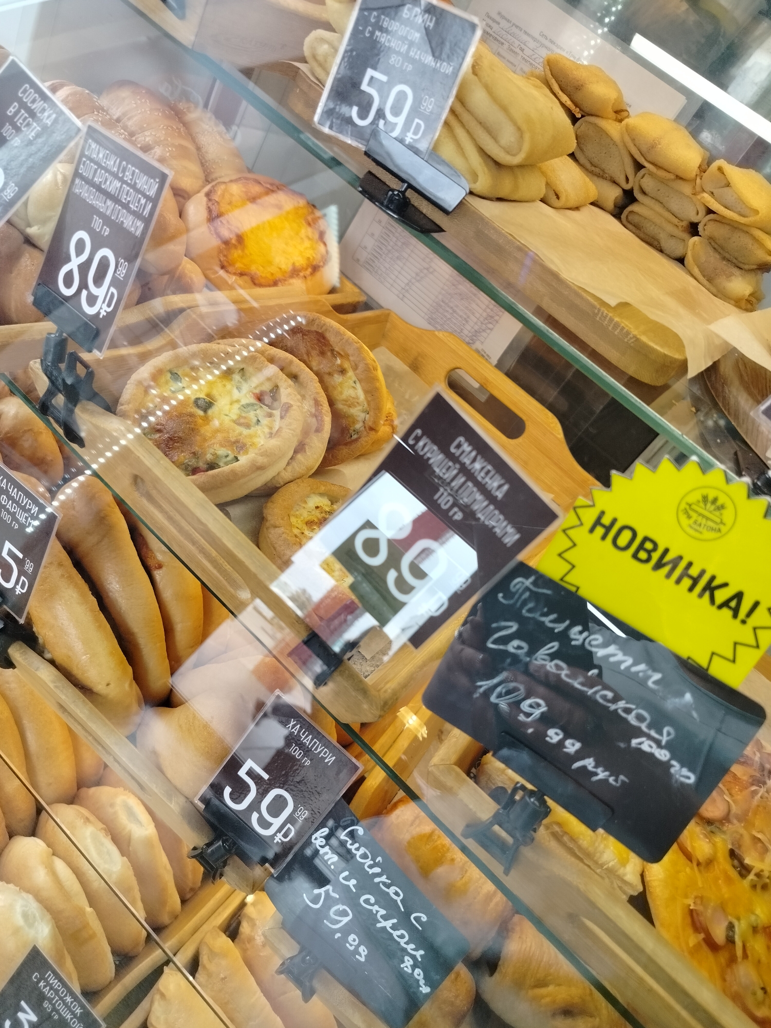 The image shows a display case filled with various baked goods, likely in a bakery or supermarket in Kemerovo, Russia. The display case is made of glass, and the lighting suggests an indoor environment. Several price tags are visible, indicating that the items are for sale.
Specific items visible include:
* **Breads and Pastries:** Various types of bread, rolls, and possibly pies or quiches are arranged on wooden shelves. Some items are topped with sesame seeds, and others appear to be filled.
* **Price Tags:** Several price tags with Cyrillic lettering and prices in rubles are displayed. Some notable tags include:
* A tag with "59.99 руб." next to a bread item.
* A tag with "89.99 руб." next to a pastry with a dark filling.
* A yellow "Новинка!" (New!) tag with a wheat logo.
* A black tag with handwritten text and "109.99 руб." indicating a new item or special offer.
* A tag for "Хачапури" (Khachapuri) with a price of "59.99 руб."
* A tag for "Пирожок с картошкой" (Small pie with potato) with a price of "85 руб." (partially visible).
The overall impression is a well-stocked bakery display ready for customers. There are no people visible in the frame. The image is taken from a slightly elevated, angled perspective, looking down into the display case. The quality of the image is good, with clear visibility of the products and text on the price tags.