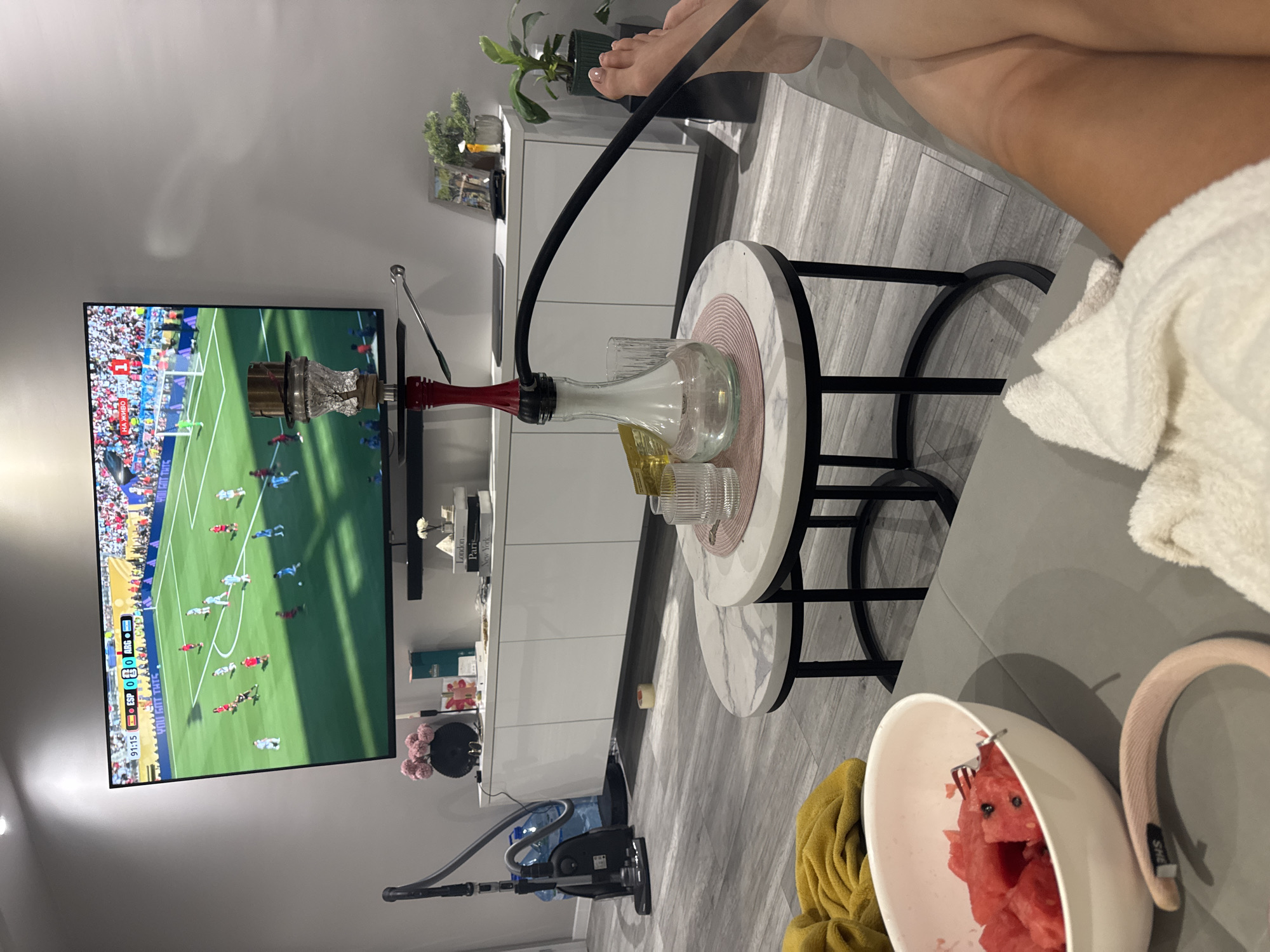 An indoor scene captured in Burgas, Bulgaria, features a person relaxing in a living room setting. A large television screen displays a live soccer match, with the score showing 0-0 between ESP and ARG at 91:15. Adjacent to the television is a hookah with smoke filling its glass base. In the foreground, a white bowl filled with chopped watermelon sits on a gray couch. A yellow blanket is draped over the back of the couch, and a white towel rests on the seat. A pair of legs, presumably belonging to the person watching the game, are visible in the lower right portion of the frame. The room's decor includes a white cabinet with books and decorative items, a vacuum cleaner, and potted plants, suggesting a modern and comfortable living space.