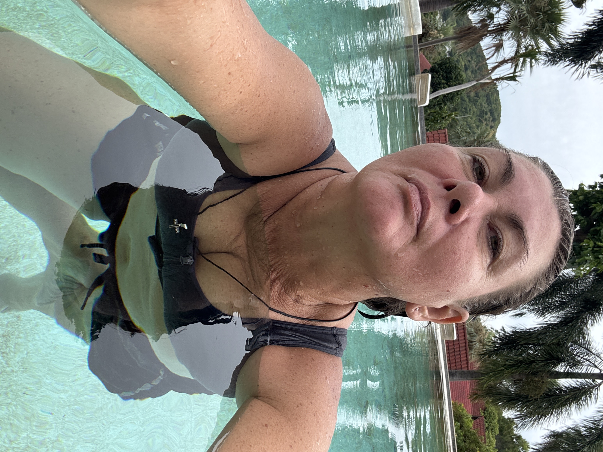 A woman is in a swimming pool, with her face and upper body visible. She is wearing a dark-colored swimsuit. The water is clear and turquoise, and sunlight is reflecting off the surface. In the background, there are palm trees and lush green hills. The sky is overcast. The location is a resort in Ban Bang Rak, Thailand.