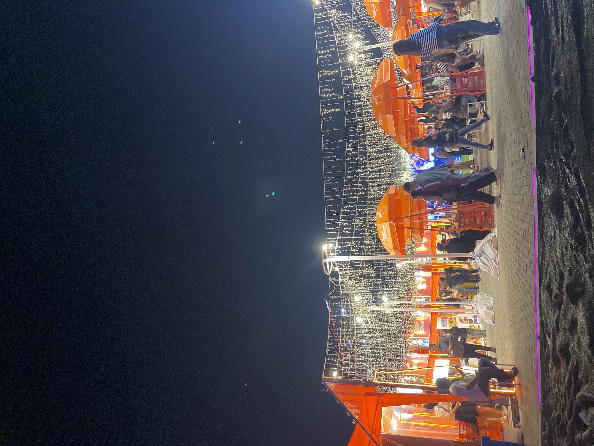 The image depicts a bustling night scene along a paved walkway in Salembaran, Indonesia. Numerous people are present, some seated at tables under orange umbrellas, others standing or walking along the path. String lights are hung overhead, illuminating the area. Food and drink vendors are visible along the side, their stalls adorned with signs. The sky above is dark. The right edge of the frame is partially obscured by dark vegetation.