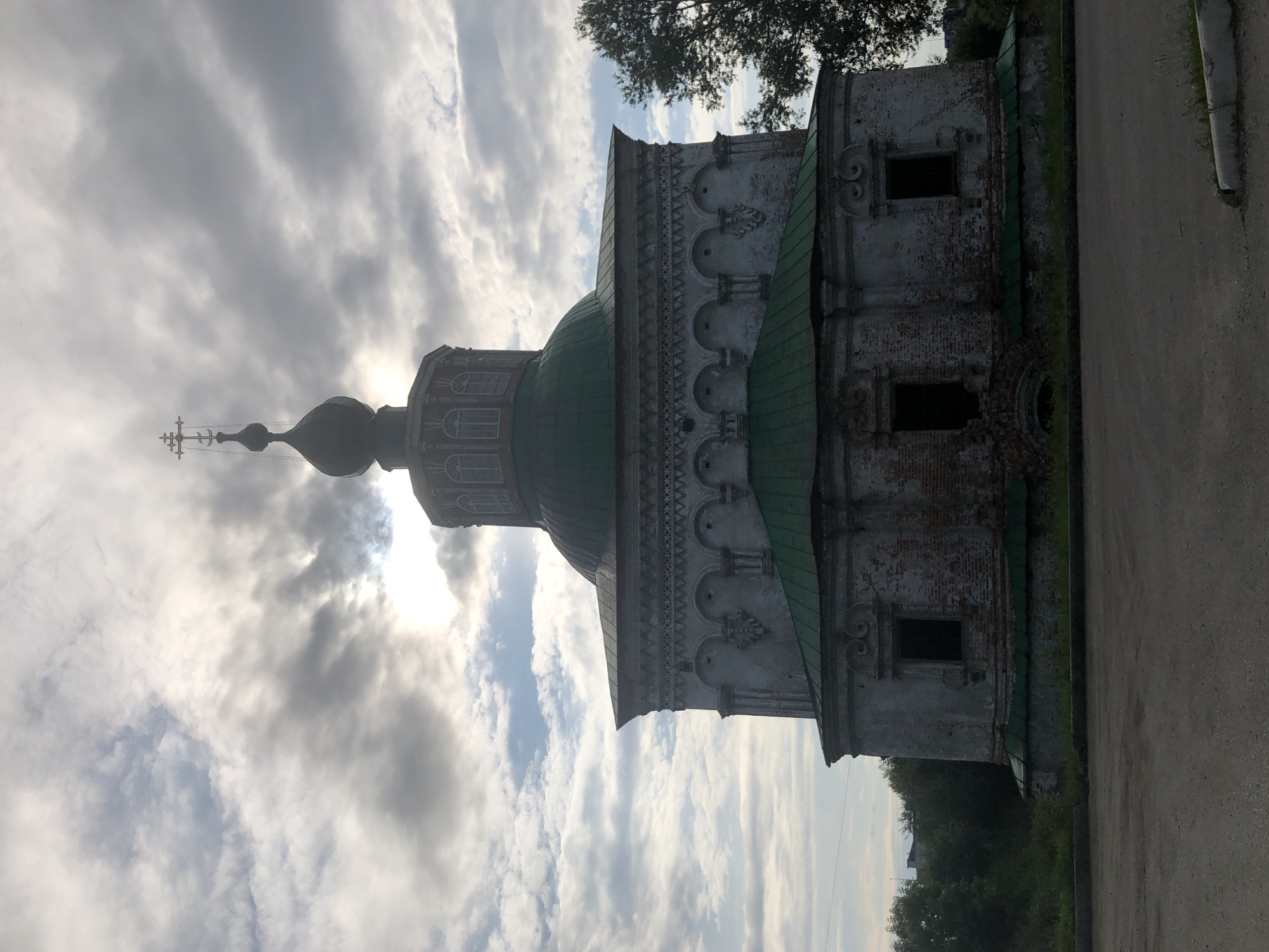 The image shows the Church of the Assumption of the Virgin, located in Solikamsk, Russia. The church is a white, brick building with a green roof and a dark dome topped with a cross. The building appears to be old and somewhat dilapidated, with visible signs of wear and tear on the brickwork. The church has several dark, rectangular windows. The building is surrounded by greenery, with trees visible on the right side and in the background. The sky is cloudy, with the sun partially visible through the clouds. The foreground is a dirt or gravel surface, possibly a parking lot or unpaved road.