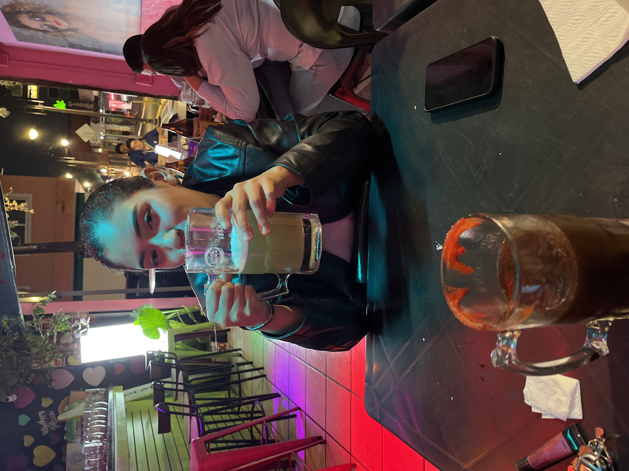 A woman in a black leather jacket is holding a glass of beer in a brightly lit establishment in San Andrés Cholula, Mexico. Another beer mug with a chili-coated rim is on the table. A cell phone, keys, and a white napkin also sit on the table. Other patrons are visible in the background.