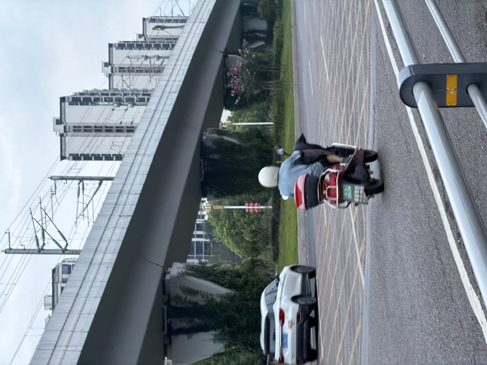 In Jiangjin, China, on a cloudy day, a person is riding a red scooter on a road under an elevated structure. The scooter rider, wearing a light blue shirt and a white helmet, is positioned in the center of the frame, facing away from the camera and traveling to the right. A white SUV is visible on the left side of the road, also traveling to the right. The elevated structure, appearing to be a bridge or overpass, casts a shadow over the road. The background includes several tall residential buildings with dark-trimmed windows, and power lines crisscross the sky above. Green trees and bushes line the side of the road. A metal guardrail with yellow reflective markers runs along the bottom of the frame.