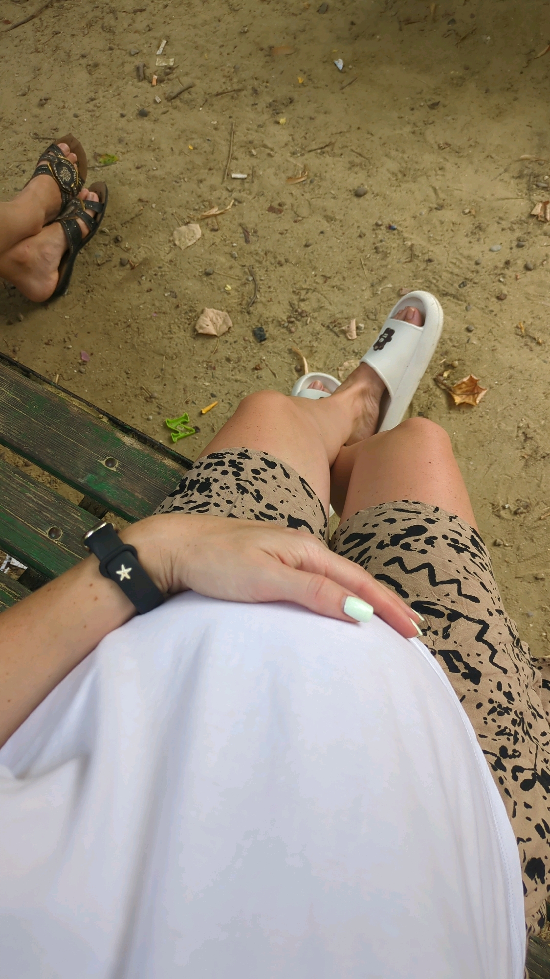 The image shows two individuals' legs and torso. The person on the left is wearing brown sandals and sitting on a wooden bench. The person on the right is wearing white slides and a leopard-print skirt, and is also sitting on the bench, with their hand resting on their stomach. The ground is covered in sand with some debris, such as cigarette butts and small pieces of wood and leaves. The image appears to be taken outdoors, possibly in a park or a recreational area. There are no clear indicators of the specific location in Chișinău, Moldova.