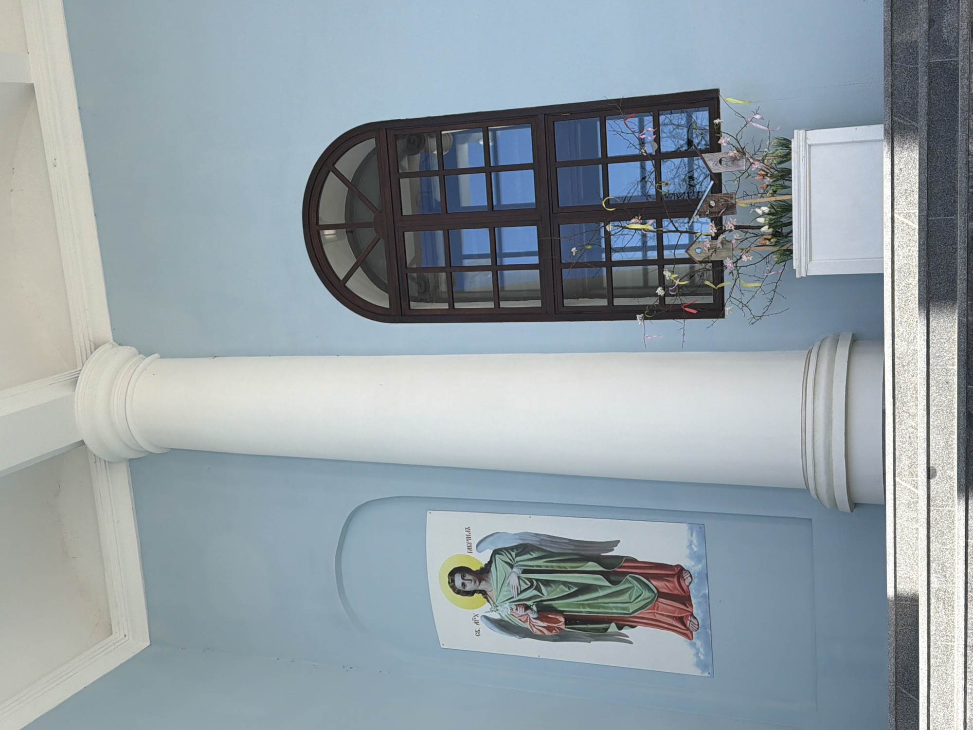 A view of the exterior of a building in Votkinsk, Russia, featuring a window with brown wooden framing and a semi-circular top. An architectural column with white trim runs vertically to the left of the window. An image of an angel is affixed to the blue wall below the column and to the right of it. There is a white rectangular planter box with branches, small birdhouses, and flowers in front of the window. A section of stairs made of gray stone runs along the right side of the scene. The building's walls are a light blue color.