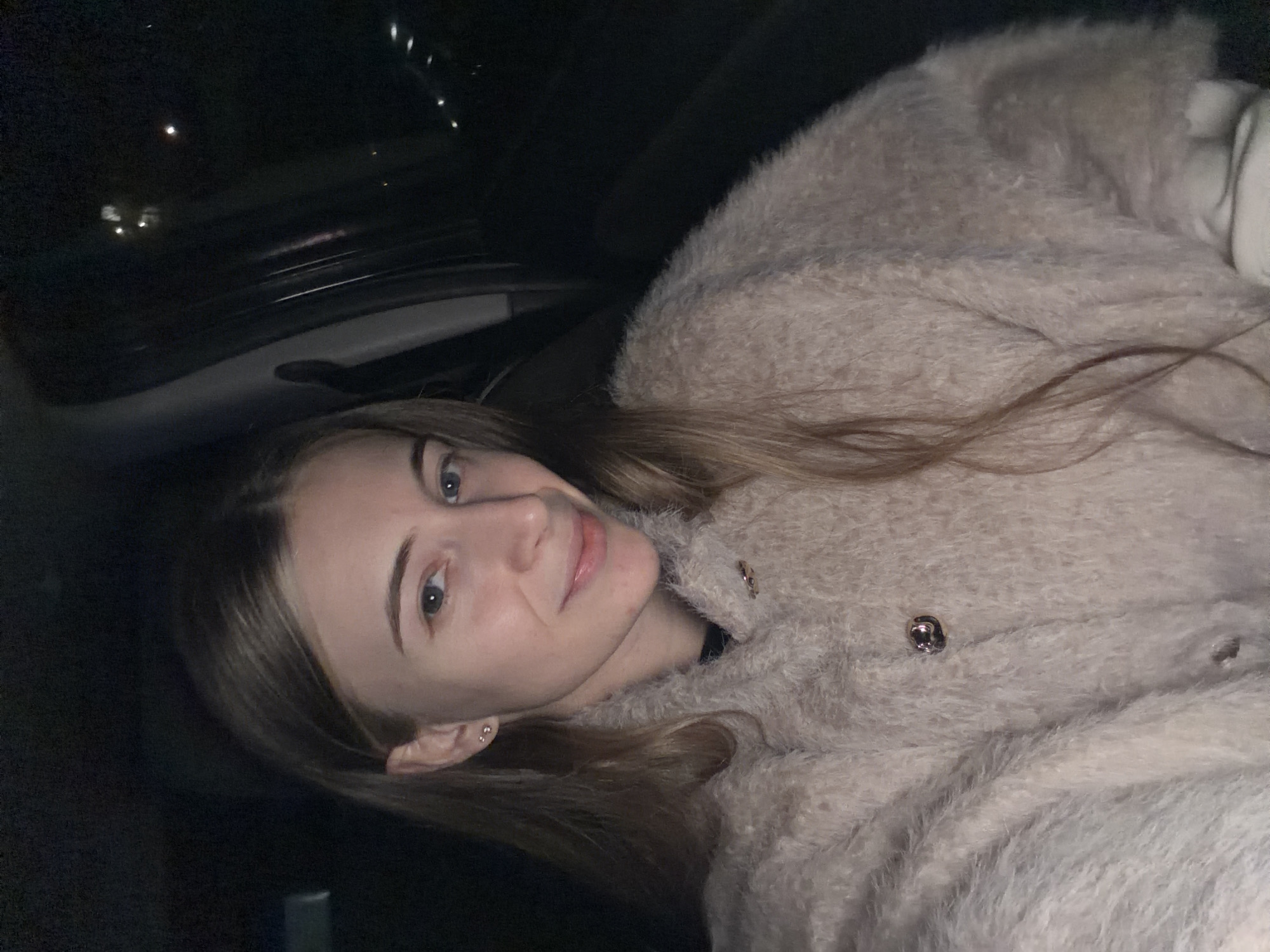 A fair-skinned female with blonde hair is the primary subject in the image. She has light blue eyes and is wearing a light-colored, textured cardigan or jacket. She is in a seated position, likely inside a car due to the presence of a car roof visible in the upper left corner. The location cannot be determined as Plovdiv, Bulgaria, as there are no indicators of location in the image. The lighting is dim, suggesting it might be nighttime.