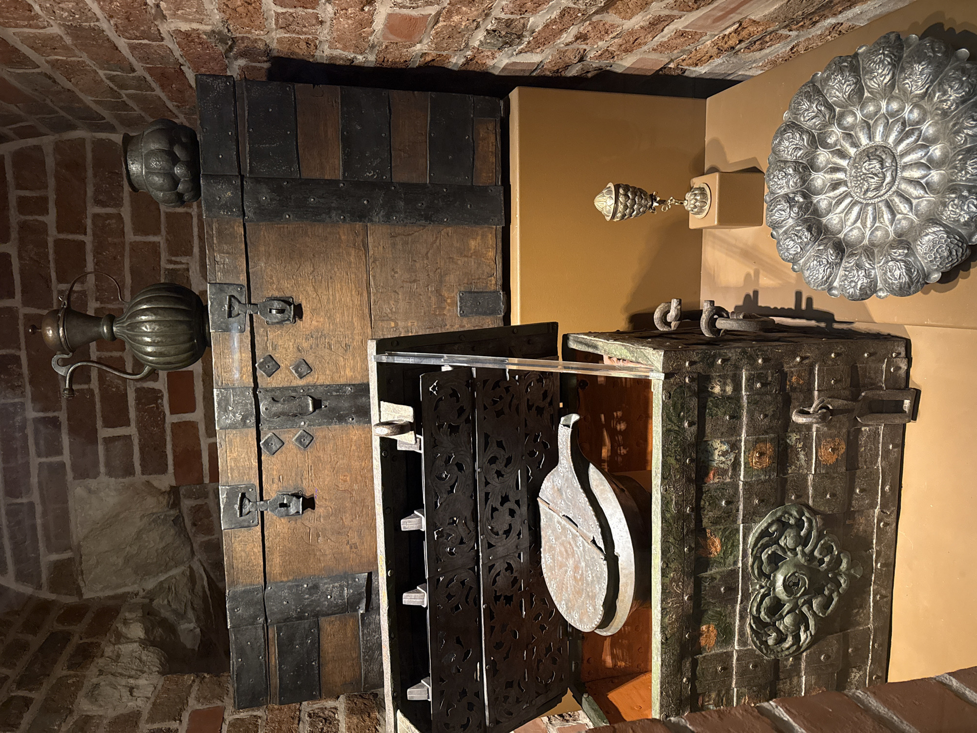 The image depicts a room within the Armoury Chamber of the Moscow Kremlin in Moscow, Russia. The room appears to be a museum display, with various historical artifacts. On the left is a tall, vertical wooden cabinet or wardrobe, reinforced with dark metal straps and fittings. Hanging next to it is a metal ewer or pitcher with a bulbous body. In the center is a chest with an intricately carved black metal door, slightly ajar, revealing an oval-shaped, two-piece object hanging inside. To the right is another chest, constructed from metal and adorned with a large decorative metal rosette. Mounted on the wall behind the chest are decorative metal objects, including a large, round plate with floral designs and a smaller, pineapple-shaped ornament. The walls of the room are made of red brick, and the lighting is somewhat dim, highlighting the textures and details of the artifacts.