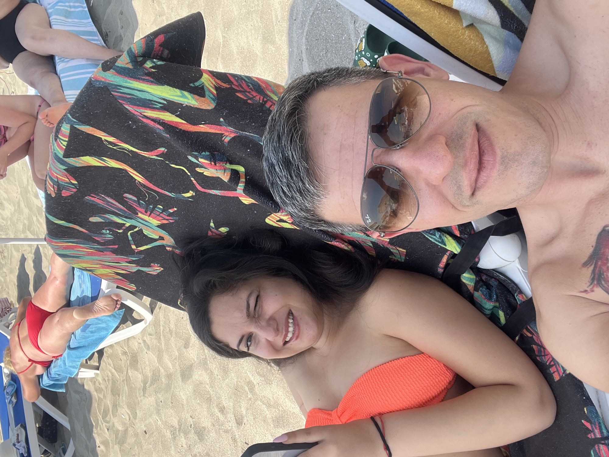 In Pomorie, Bulgaria, two individuals are captured in a selfie on a sandy beach. The woman, positioned on the left, lies on a sun lounger, her face turned towards the camera with one eye winking and a smile. She wears an orange bikini top and a black bracelet. Beside her, on the right, a man wearing aviator sunglasses and a grey and white striped towel is also visible. His chest displays a tattoo. In the background, a woman in a red bikini relaxes on another sun lounger. The beach is composed of fine sand, and the overall setting suggests a casual day at the seaside.