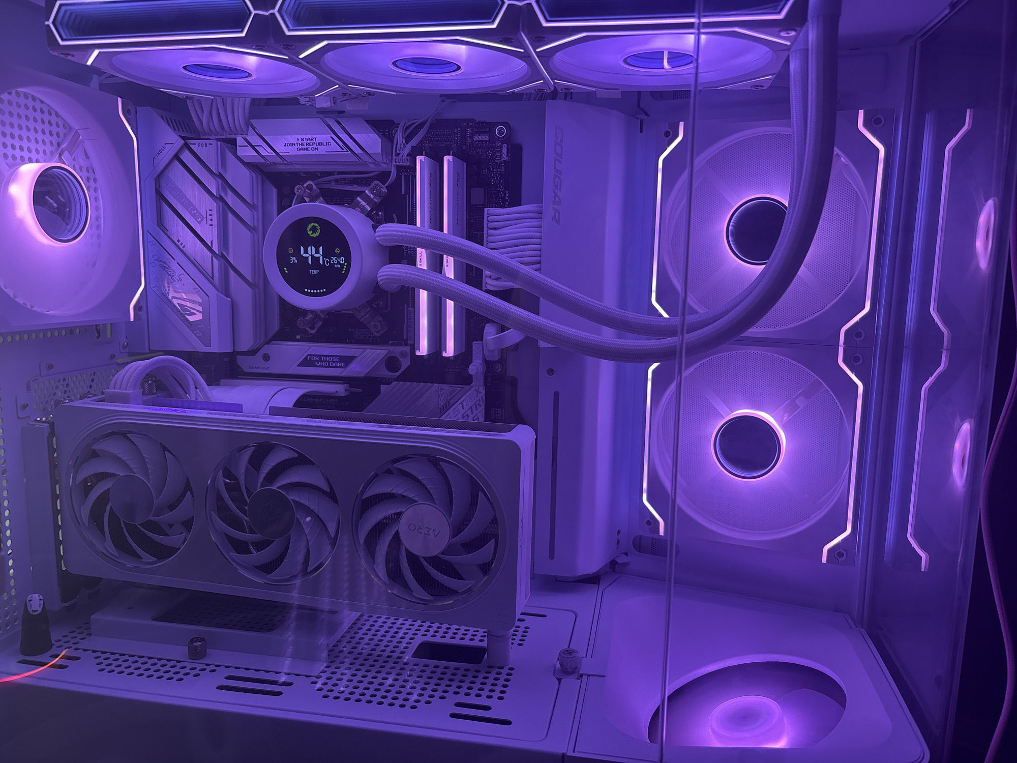 This is a close-up, interior shot of a white custom-built PC case, illuminated by purple RGB lighting. The case is clean and organized, showcasing a high-end gaming setup.
Prominent components visible include:
* **Motherboard:** A white ASUS ROG Strix motherboard with heatsinks and M.2 covers.
* **CPU Cooler:** An AIO liquid cooler with a circular digital display showing 44°C and 2640 RPM. The tubes are white and connect to the CPU block.
* **RAM:** Two sticks of white DDR4 RAM are installed, with pink RGB lighting.
* **Graphics Card:** A white ASUS ROG Strix RTX graphics card with three large fans. The "AERO" branding is visible on the shroud.
* **Fans:** Multiple RGB fans are present. Two large fans are mounted vertically on the right side of the case, and two more are visible at the top, also emitting purple light. A fan is visible at the bottom of the case, also lit in purple.
* **PSU Shroud:** A white shroud covers the power supply unit.
* **Case:** The PC case itself is white, with a clear side panel allowing visibility of the internal components. The interior is well-lit with RGB accents.
A small, dark figurine resembling "No-Face" from Spirited Away is placed on a shelf below the graphics card, adding a personal touch to the build. The overall aesthetic is modern, clean, and designed for performance and visual appeal, typical of a enthusiast PC build. The context suggests the image was taken indoors, likely in a home environment, focusing solely on the computer hardware.