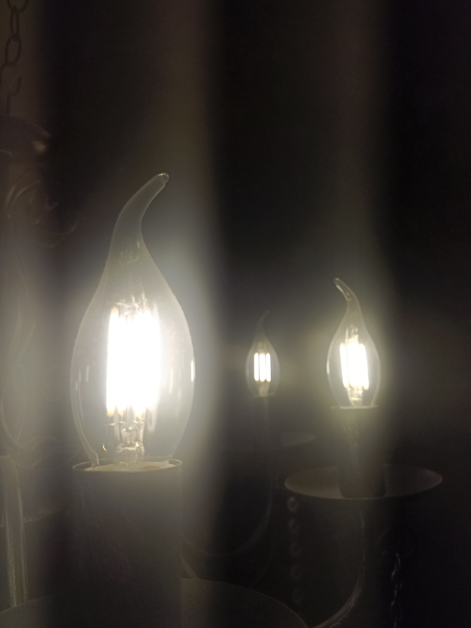 The image shows a close-up of three candelabra-style LED light bulbs, each with a clear glass flame-shaped outer casing and internal light-emitting diodes. The bulbs are illuminated and emit a bright white light. The leftmost bulb is the largest and most prominent, positioned in the foreground. Two smaller bulbs are visible in the background, slightly to the right and further back. The bulbs are mounted on dark, ornate fixtures that are partially visible. The overall lighting is dim, with the light from the bulbs creating a hazy glow and casting soft shadows. The background is dark and out of focus, suggesting a dimly lit interior environment. There are no people or animals in the image, and no discernible Moscow landmarks or specific locations. The scene appears to be an interior decorative lighting fixture.
