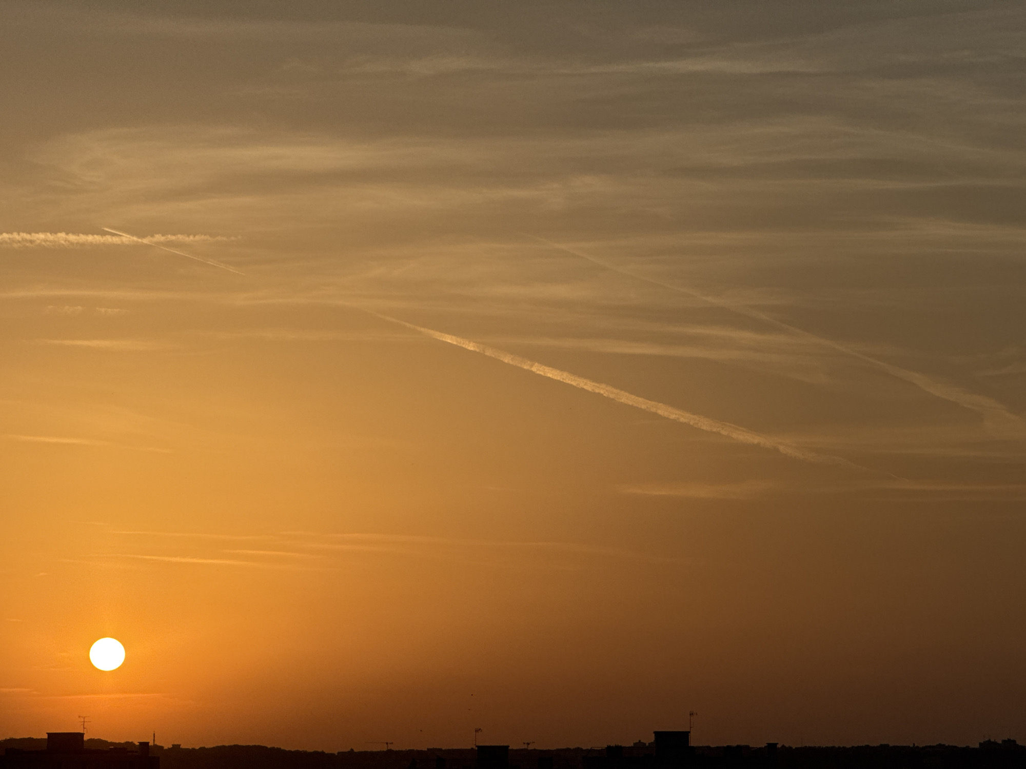 A wide shot captures a sunset over a silhouetted cityscape. The sun, a bright white orb, sits low on the left horizon, casting a warm orange glow across the sky. Wispy clouds and contrails stretch across the sky in various directions. The foreground is dominated by the dark silhouettes of buildings, some with antennas or cranes, against the vibrant sky. The scene is devoid of people and focuses solely on the natural beauty of the sunset and the urban landscape it illuminates.