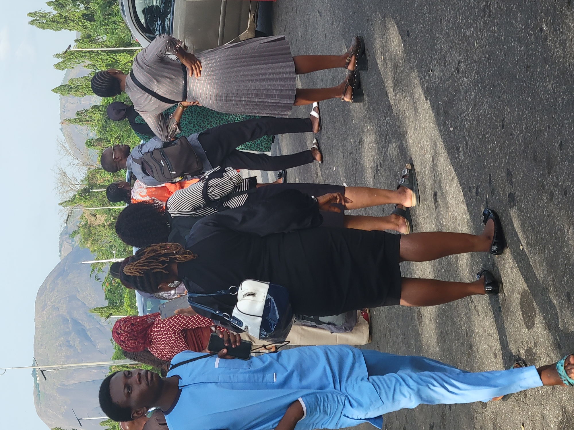 The image shows a group of people standing on a road in what appears to be a semi-urban or suburban area, possibly in Abuja, Nigeria. A large rock formation is visible in the background, suggesting a location on the outskirts of the city where such geological features are common. The people appear to be waiting, possibly for transportation or an event. They are dressed in a variety of attire, including formal and casual clothing. Some are holding phones or bags. The road is paved and shows signs of wear. A vehicle is partially visible in the upper portion of the image.