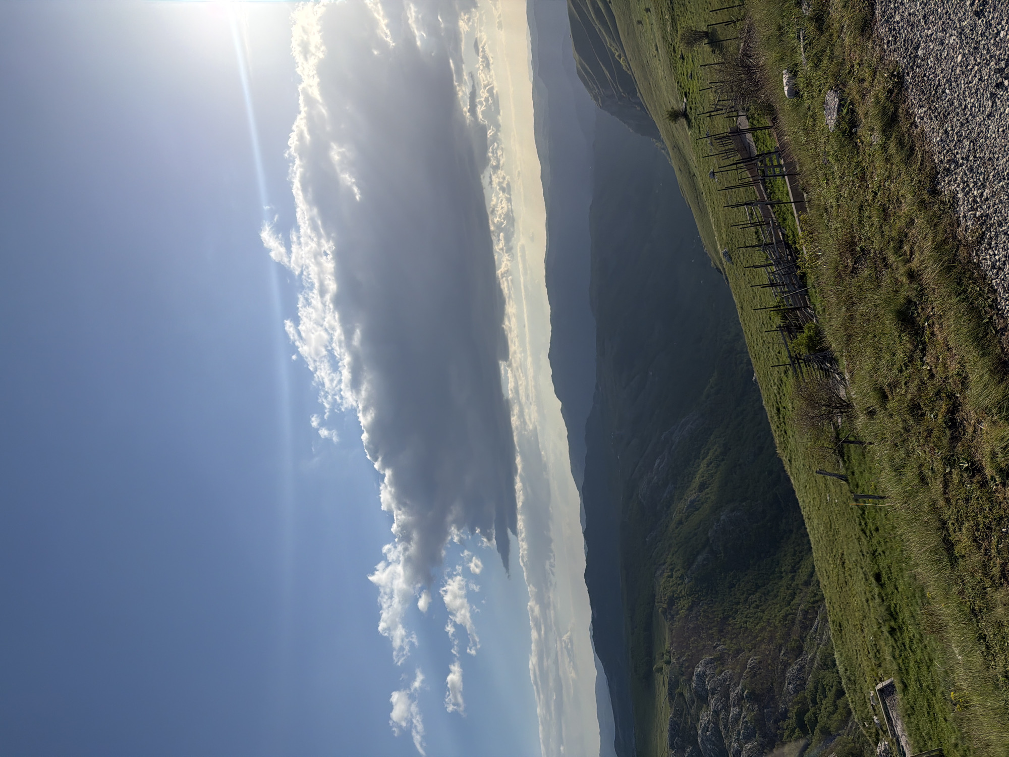 The image depicts a landscape near Plužine, Montenegro. The sky is blue with white clouds and a bright sun. A range of mountains extends into the distance. The foreground features a grassy hillside with a wooden fence and gravel.
