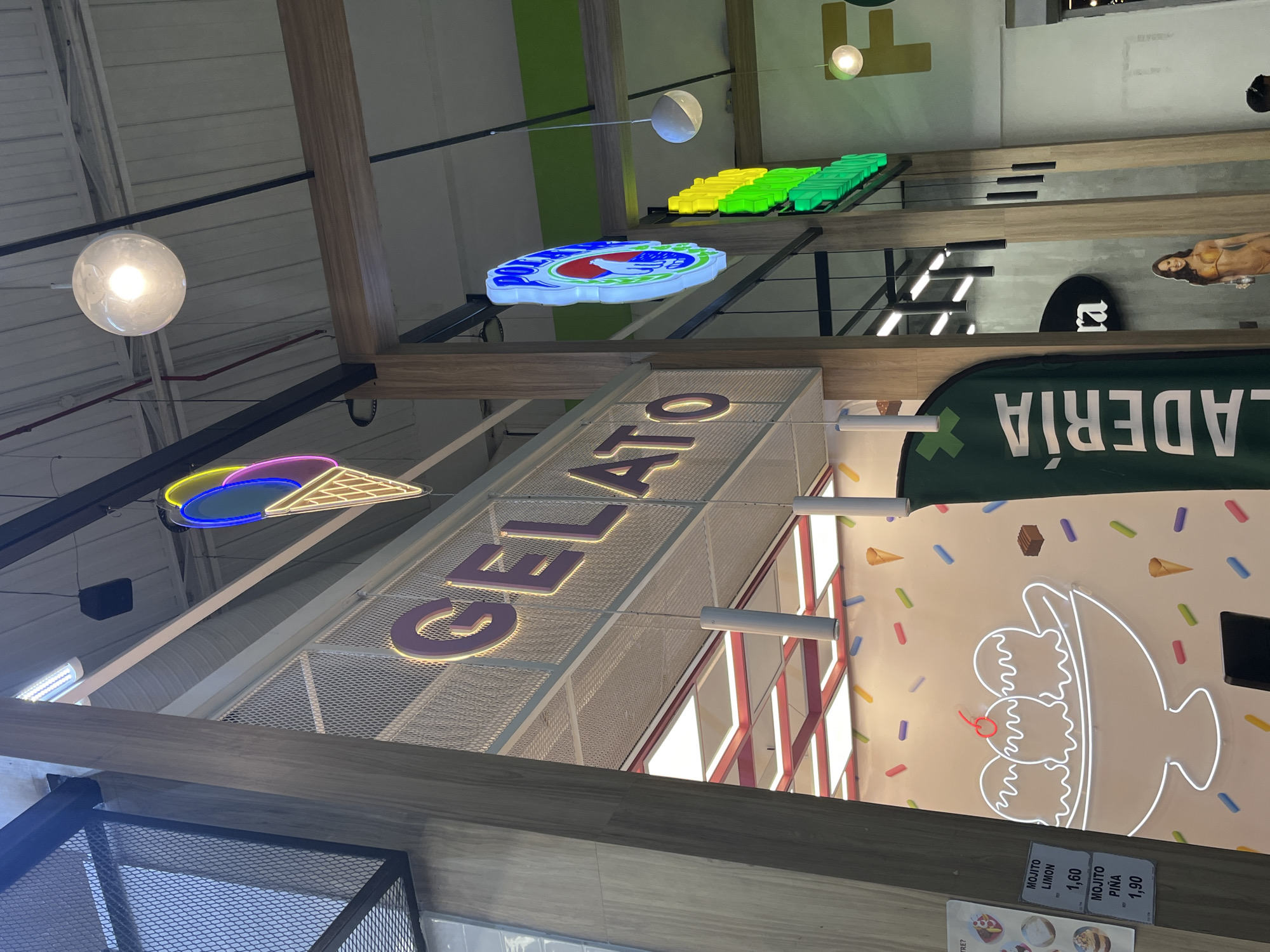 The image depicts the interior of a gelato shop in Porlamar, Venezuela. The shot is taken from a low angle, looking up towards the ceiling where various signs are suspended. The signs include "Gelato" in large, illuminated letters, an ice cream cone outline in neon lights, and a green banner with the word "Heladería" (ice cream shop in Spanish) visible. There's also a sign in blue and white that says something like "Polo Norte" that is partially obstructed. There are ice cream themed decorations on the walls, including a neon ice cream sundae. The lighting is a mix of natural and artificial light, with hanging spherical lamps. The shop appears to be located in a commercial area, evidenced by the various signs and advertisements. The style of the shop is modern and visually appealing.