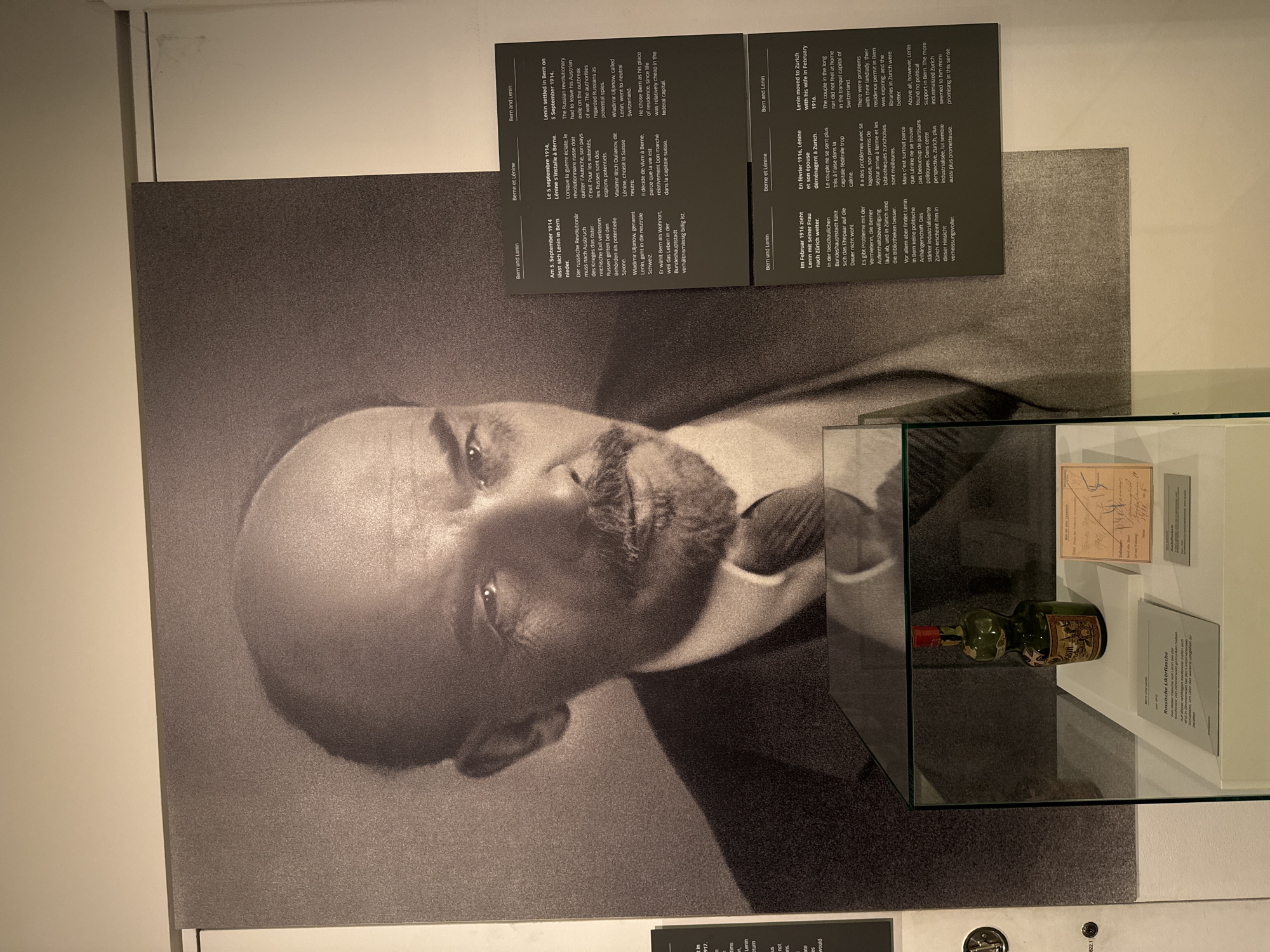 This media file is a photograph of a museum exhibit. The primary subject is a large black and white portrait of Vladimir Lenin, with his face angled and his gaze directed to the left. To the right of the portrait is a glass display case containing a small, green glass bottle with a red cap, and several documents. Behind the bottle and documents, a small framed text is visible.
In the upper right corner of the image, there are three rectangular plaques with text, likely providing information about the exhibit. The text on the plaques is visible and appears to be in at least three languages: German, French, and English. These plaques detail Lenin's time in Bern and Zurich, Switzerland, in 1914-1916.
The scene is indoors, likely in a museum or exhibition space, with plain walls and lighting that illuminates the display. The overall impression is one of historical documentation and preservation. The bottle and documents in the display case suggest personal effects or artifacts related to Lenin's life during his exile.