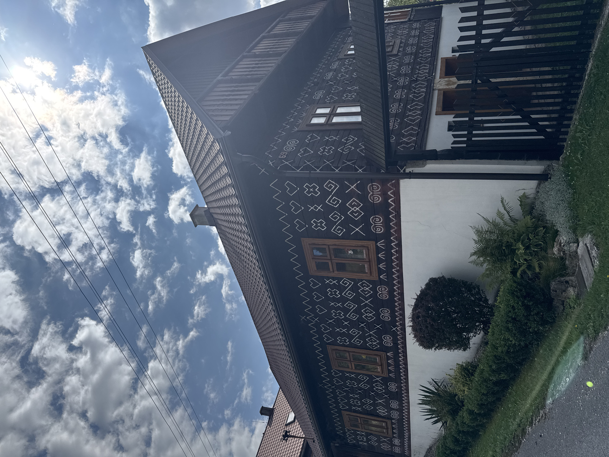 The image shows a traditional wooden house in the village of Čičmany, Slovakia, known for its unique white geometric patterns painted on dark wooden facades. The house has a dark, gabled roof and features several windows with wooden frames. The upper part of the house is adorned with intricate white folk art designs, including zigzags, hearts, crosses, and spiral motifs. A wooden balcony runs along the upper story, supported by wooden posts. In front of the house, there is a white-washed lower story, a black wooden fence, and lush green vegetation, including bushes, ferns, and a small tree. The sky above is blue with scattered white clouds, and sunlight filters through, creating a bright atmosphere. Electrical wires are visible strung across the sky in the upper left portion of the image. The scene captures the distinctive architectural style characteristic of Čičmany.