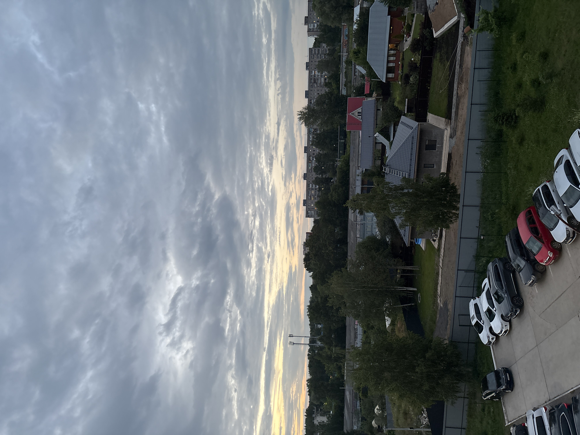 The image depicts a panoramic view of a suburban area in Chaikovsky, Russia, under a cloudy sky during twilight. In the foreground, a parking lot contains several parked cars, including a black Kia, a white SUV, a white sedan, a dark gray SUV, a black SUV, and a red hatchback. Behind the parking lot, a fence separates it from a green grassy area and several houses with gray roofs. Further in the background, a dense line of trees separates the houses from a row of tall, gray apartment buildings. Two tall radio towers are visible in the distance, positioned above the tree line, and the sky displays streaks of orange and yellow light, indicating the setting sun. The overall scene suggests a residential neighborhood on the outskirts of a city.