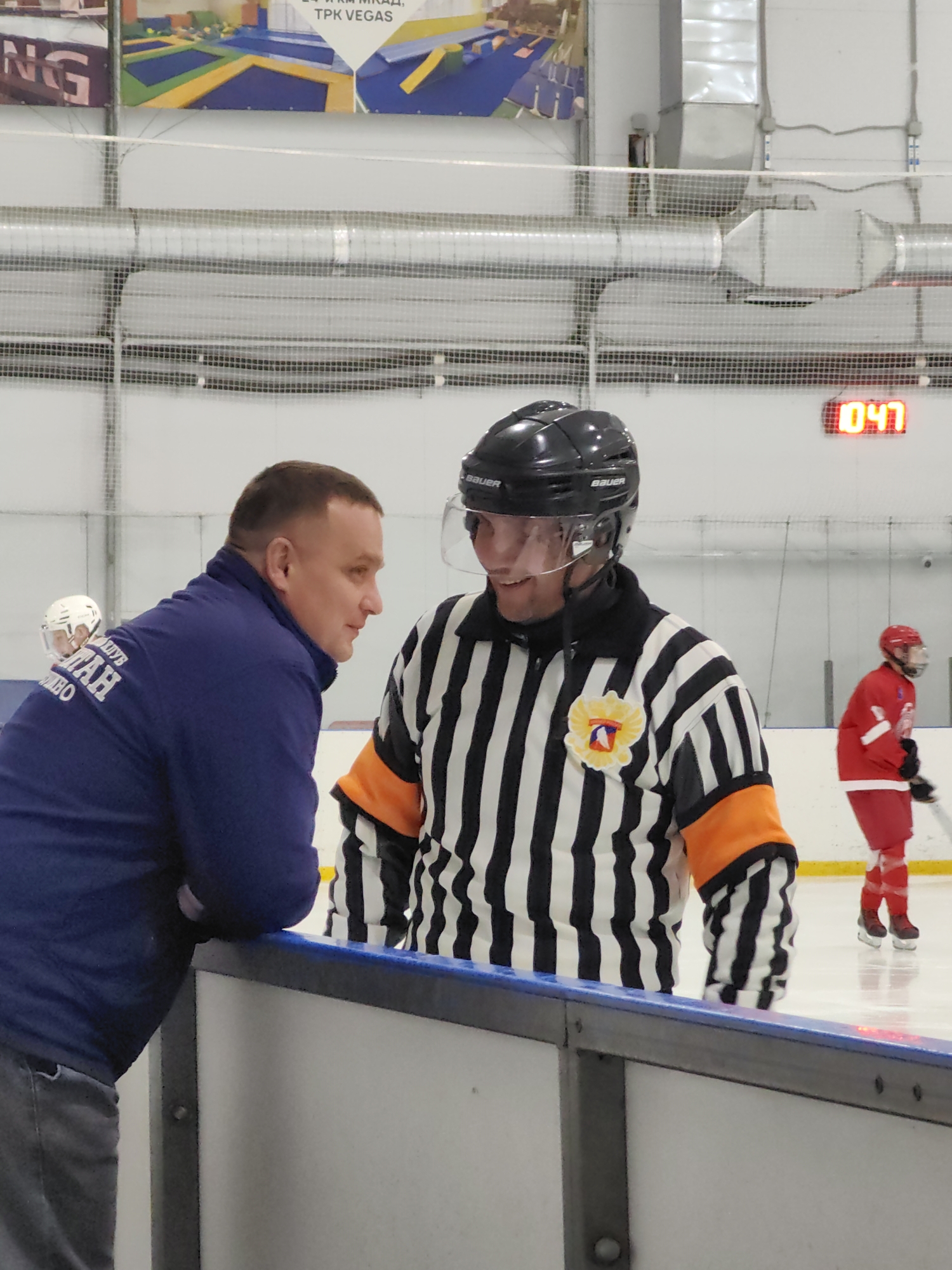 Two men are talking on an ice rink. The man on the left is wearing a dark blue jacket and grey pants, and is leaning on the boards of the rink. The man on the right is wearing a referee's uniform with black and white stripes, an orange band on his left sleeve, and a black helmet. He is smiling and appears to be talking to the man on the left. In the background, a person in a red hockey uniform is visible on the ice, and a digital clock on the wall displays "0:47". The environment appears to be an indoor ice rink, possibly part of the "TPK VEGAS" complex visible in the background, which is located in the city of Vidnoye, Russia.