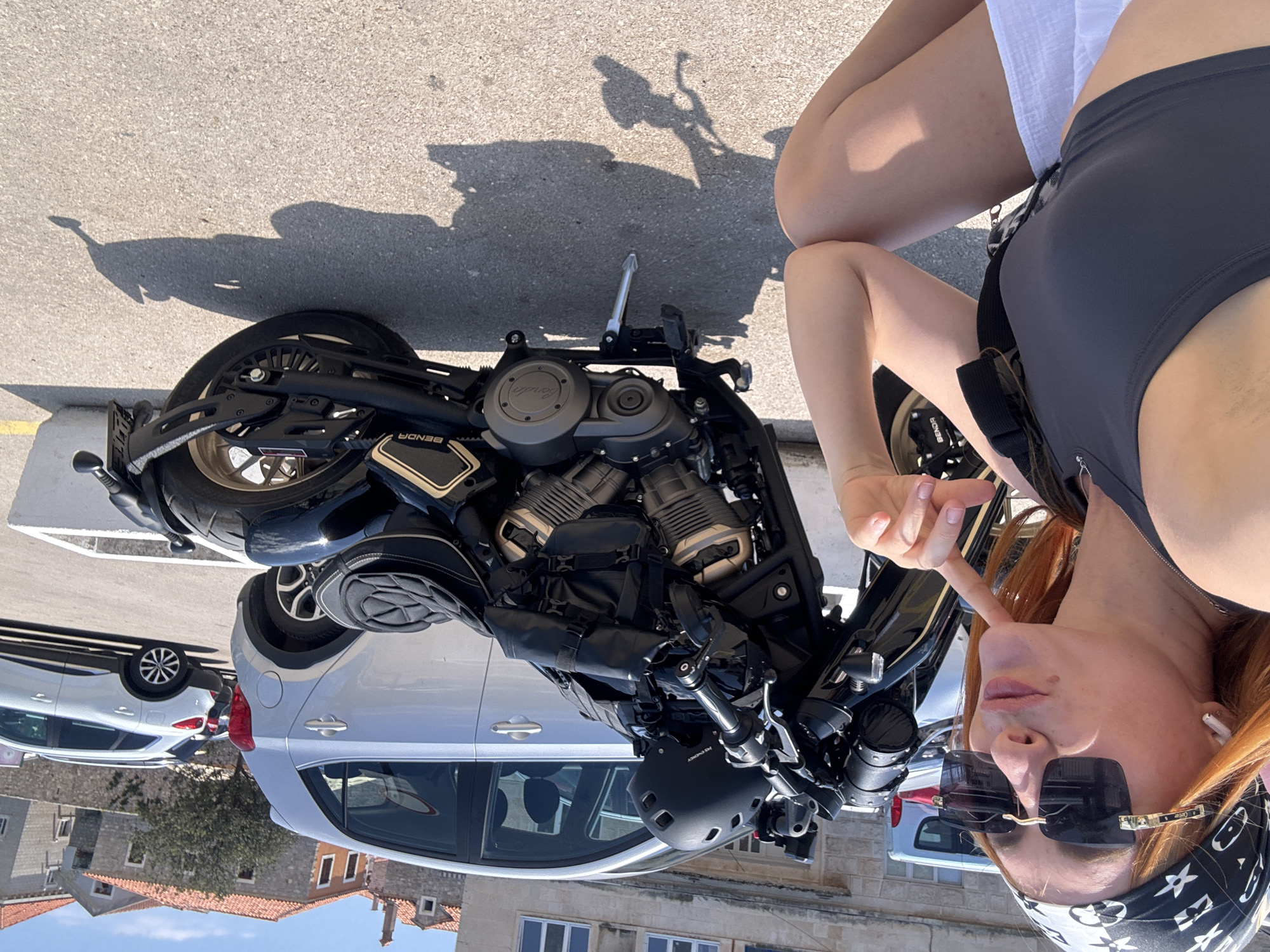 The media file is an outdoor, eye-level, slightly low-angle shot taken on a sunny day. The scene depicts a woman in the foreground, with her upper body and head visible, wearing sunglasses and a black bandana. She is touching her face with her right hand, and her left hand is near the handlebars of a black motorcycle. The motorcycle is the central object in the middle ground, partially obscuring a silver car behind it. Buildings with red tile roofs and stone facades are visible in the background, suggesting a Mediterranean or European setting. The woman's attire, including dark athletic shorts and a white top, indicates a casual or leisure activity. The bright sunlight creates distinct shadows on the asphalt ground, and the overall ambiance suggests a warm, summer day. No visible text is present on the subjects or objects, except for a logo on the motorcycle that appears to read "Benda". The image appears to be taken in Budva, Montenegro, given the provided location context.