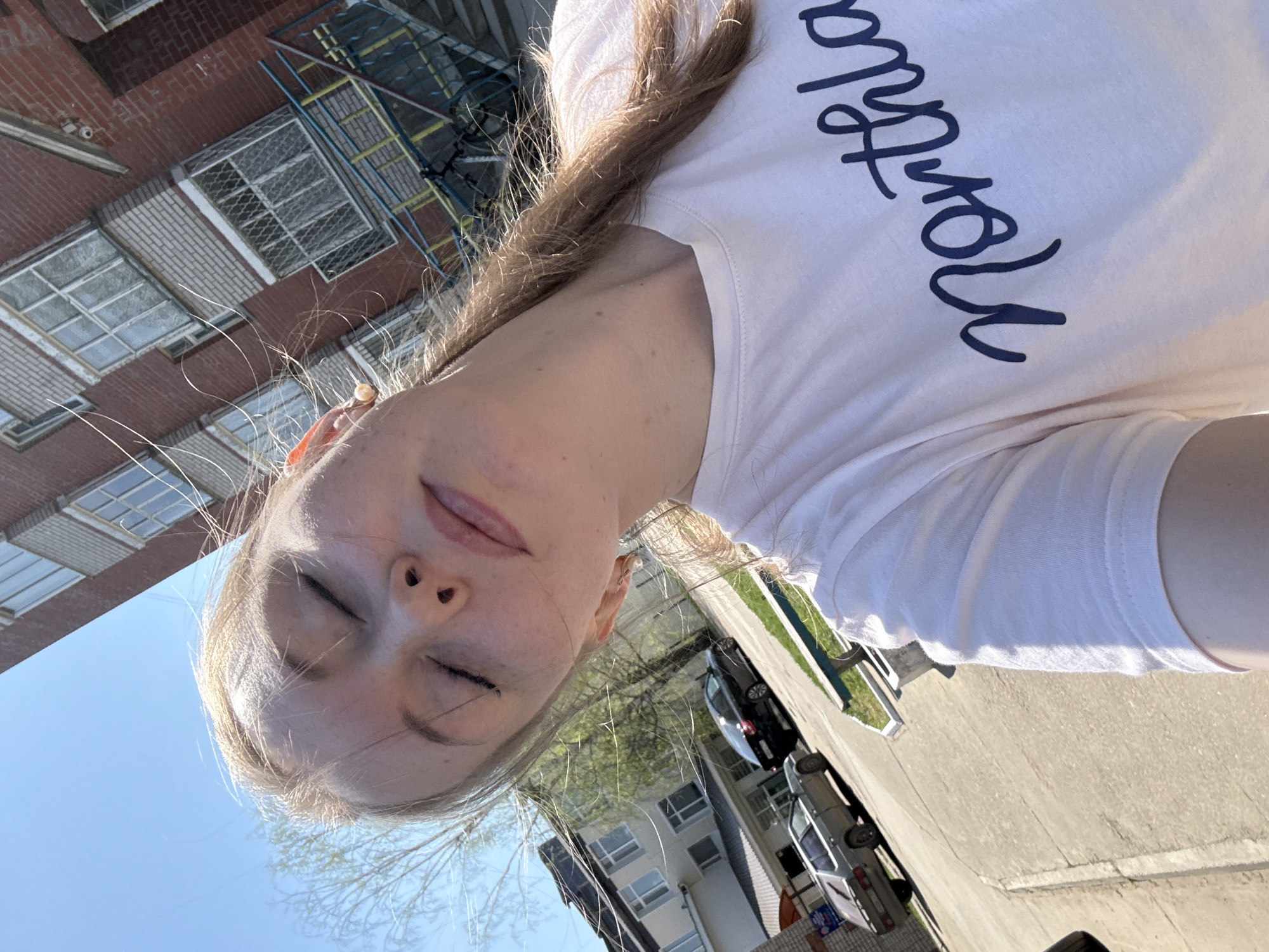 A vertically oriented photograph depicts a fair-skinned individual wearing a white t-shirt with the word "Northland" printed on it in blue cursive script. The person has blonde hair and closed eyes, facing the camera at an odd angle. The background shows an urban scene with buildings constructed from brown brick and white brick, complete with windows. A vehicle, a black car, can be seen along a gray road. Part of the clear blue sky is visible in the background.