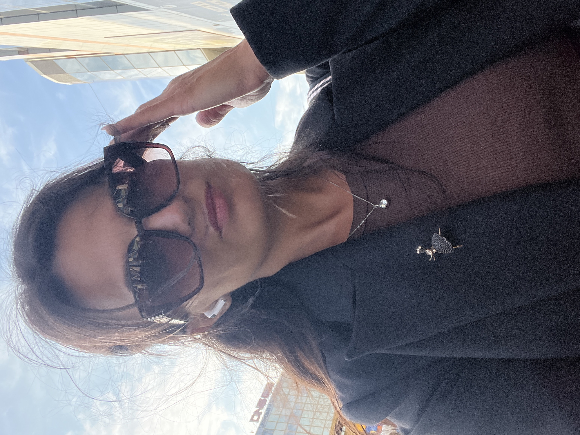 A woman is taking a selfie outdoors with a building and the sky in the background. She has long, dark hair, is wearing large, dark sunglasses, and has white earbuds in her ears. She is wearing a dark jacket over a brown ribbed top and a delicate silver necklace with a small pendant. A small, dark brooch shaped like a ballet dancer is pinned to her jacket. Her right hand is raised, adjusting her sunglasses. The environment suggests an urban setting, with modern architecture visible. This scene is not identifiable as Petropavlovsk-Kamchatsky, Russia.