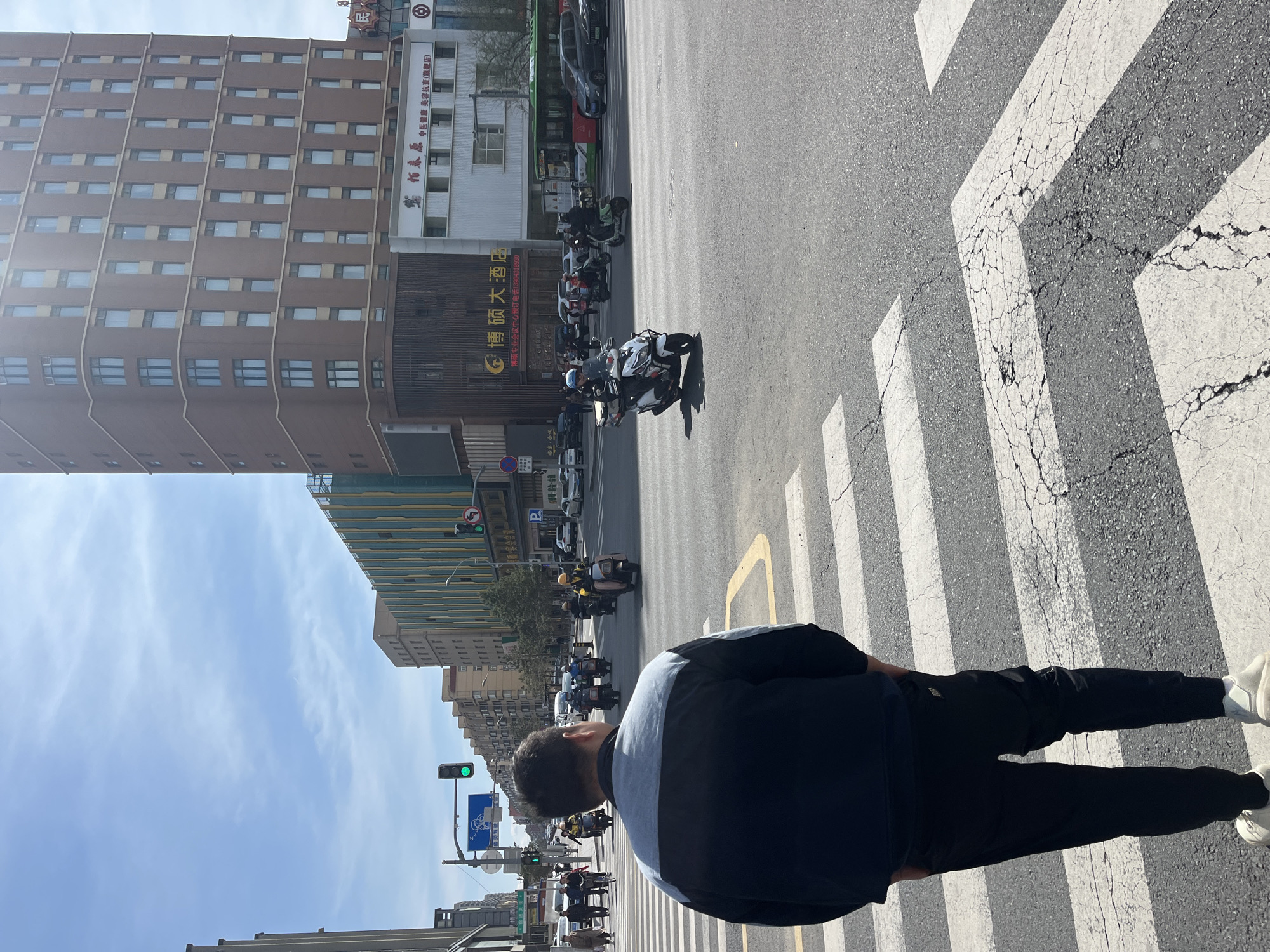 A street in Changchun City, China, is depicted in a ground-level, slightly rotated, full shot photograph. In the foreground, a man is captured from the waist down, positioned on a crosswalk marked by white stripes. The man is wearing a dark jacket with a grey stripe down the back, black pants, and white shoes.
Behind the man, the street scene unfolds with motorbikes and cars moving in the same direction. Buildings line the street, including a high-rise brick building on the left and a building with Chinese characters on the facade on the right. Traffic lights are visible in the background. The sky is light blue with sparse clouds.
