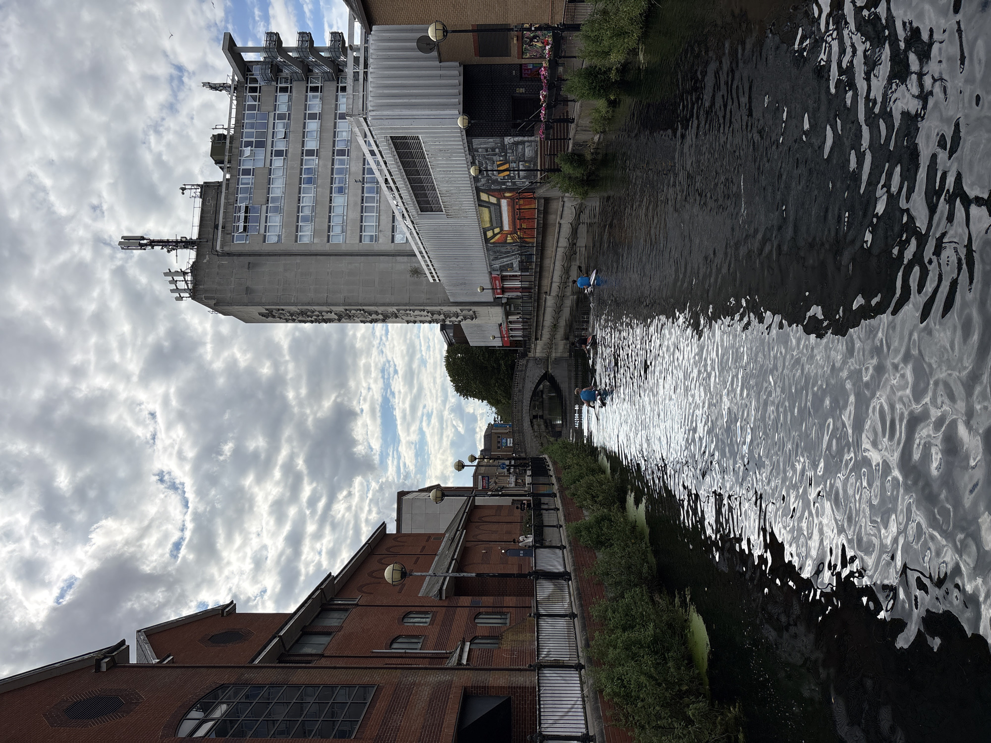 The image depicts a canal with buildings on either side. On the left, a tall, modern grey building with many windows dominates the scene, featuring what appears to be telecommunications equipment on its roof. In front of it, a red brick building with arched windows and a sloped roof is visible. On the right side of the canal, more brick buildings line the waterway. The canal itself is dark and reflects the sky and surrounding structures. Greenery is present along the banks. In the distance, a stone bridge arches over the canal.
A person is visible in the canal, seated in a small boat, paddling forward. The sky is filled with dramatic, white, and grey clouds, suggesting a dynamic weather condition. The lighting suggests it is daytime, possibly late afternoon or early evening due to the contrast and shadows. The scene has an urban feel with a mix of modern and older architecture alongside the natural element of the canal. No visible text is discernible in the image.