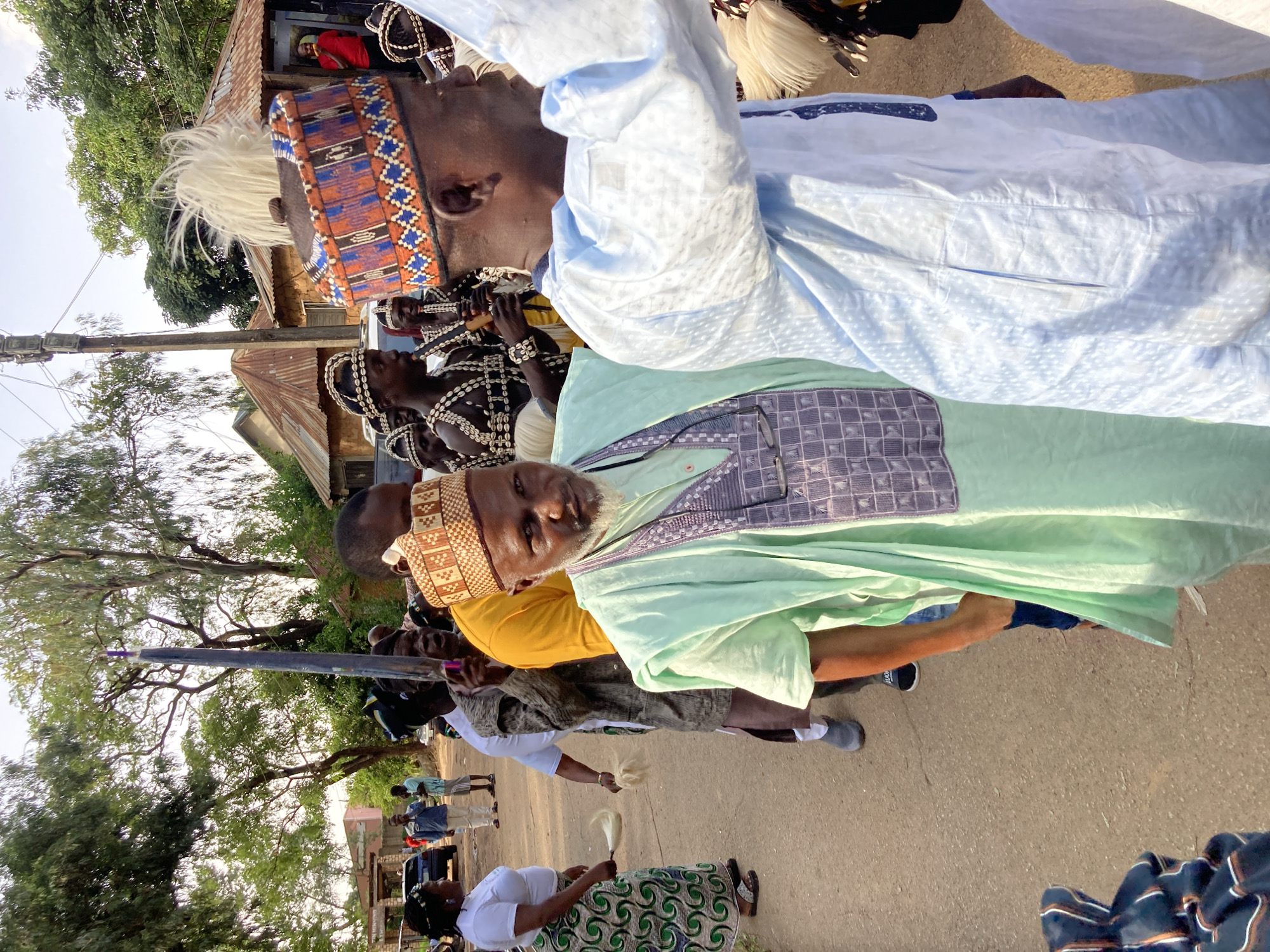 In Jos, Nigeria, a group of men are participating in a procession along a dusty, paved road, possibly for a cultural event or ceremony. The men are dressed in traditional attire, including colorful hats adorned with beads and intricate patterns, and flowing robes. Some are wearing necklaces made of shells or beads. In the foreground, a man in a light green robe with a patterned bib is looking towards the camera, with a grey beard. To his right, another man in a light blue robe is facing away from the camera. In the background, several other men are visible, some carrying long, dark staffs, and one appears to be playing a musical instrument. Trees with green foliage line the sides of the road, and small buildings with corrugated iron roofs are visible in the distance. The sky is clear and bright, indicating daytime.