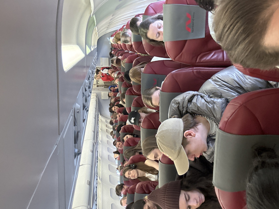 The image displays the interior cabin of a Nordwind Airlines commercial aircraft, with the indicated location being the city of Novogorelovo, Russia. The perspective is from a passenger seat, looking down the aisle towards the front and partially towards the rear. The cabin is brightly illuminated, with external daylight visible through windows on the left side of the aircraft.
Dozens of passengers occupy the dark red-upholstered seats, each featuring a gray headrest cover with the white "Nordwind Airlines" logo. Most passengers appear to be young adults, dressed in casual attire. In the immediate foreground, a male passenger wears a beige baseball cap, a shiny silver puffer jacket, and a silver chain around his neck; he is looking left with his mouth slightly open. To his left, a female passenger wears a dark brown beanie and dark clothing, looking downwards. Further back, a female passenger with long hair and white over-ear headphones has her eyes closed. Another male passenger in the mid-ground wears a black baseball cap with a white Stüssy logo. Passengers are mostly seated, some looking forward, others downwards, and some appear to be resting.
In the background, near the rear-right of the visible cabin, two female flight attendants are standing. They are dressed in red uniforms, with one blonde-haired attendant clearly visible, suggesting active duty or preparation. The overhead bins are closed and light grey, extending along both sides of the cabin.
