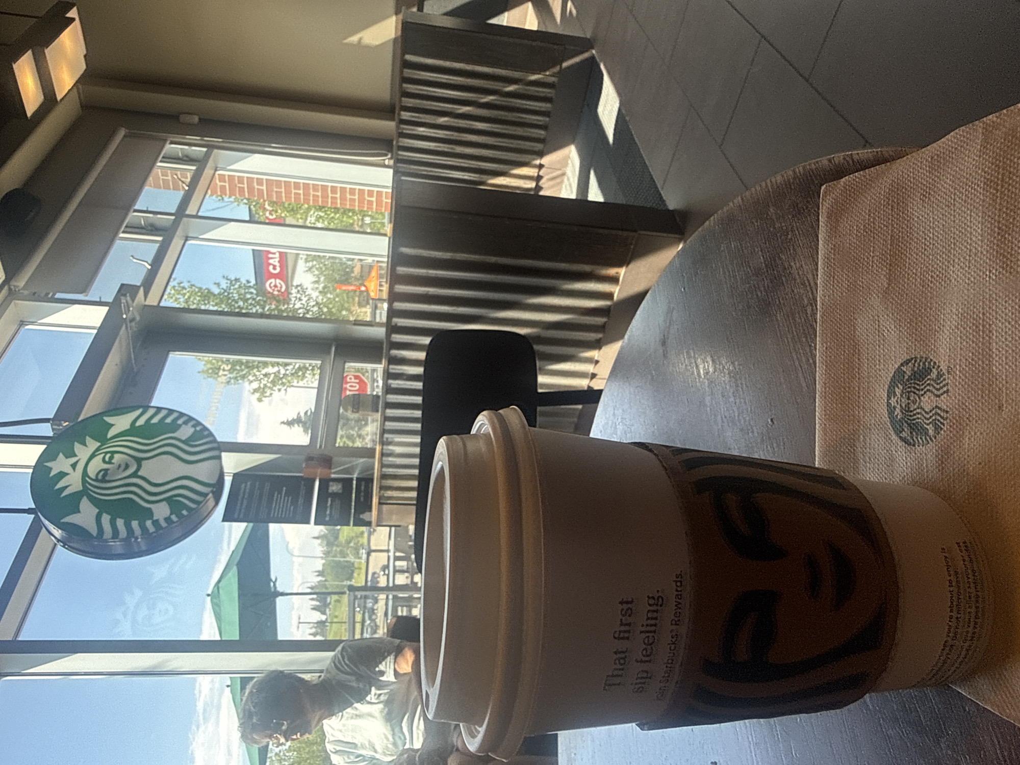 The image depicts the interior of a Starbucks cafe, with a close-up view of a disposable coffee cup with a cardboard sleeve. The cup is light brown and has text printed on it that reads "That first sip feeling. Join Starbucks? Rewards." The cardboard sleeve features the iconic Starbucks siren logo. To the right of the cup, a light brown paper napkin with a smaller Starbucks logo is visible. The scene appears to be set during daylight, with sunlight streaming through a large window. Outside the window, trees and a CALI (likely a gas station sign) are visible, along with a green umbrella. In the background, to the left, a person wearing a light-colored shirt and sunglasses is seated, looking to the left. The cafe interior features booth-like seating with corrugated metal dividers and a dark-tiled floor. The image was taken inside a Starbucks cafe in Calgary, Canada, as suggested by the presence of a CALI sign, which is a common gas station chain in Western Canada, and the overall aesthetic fitting for a Starbucks location in the region.