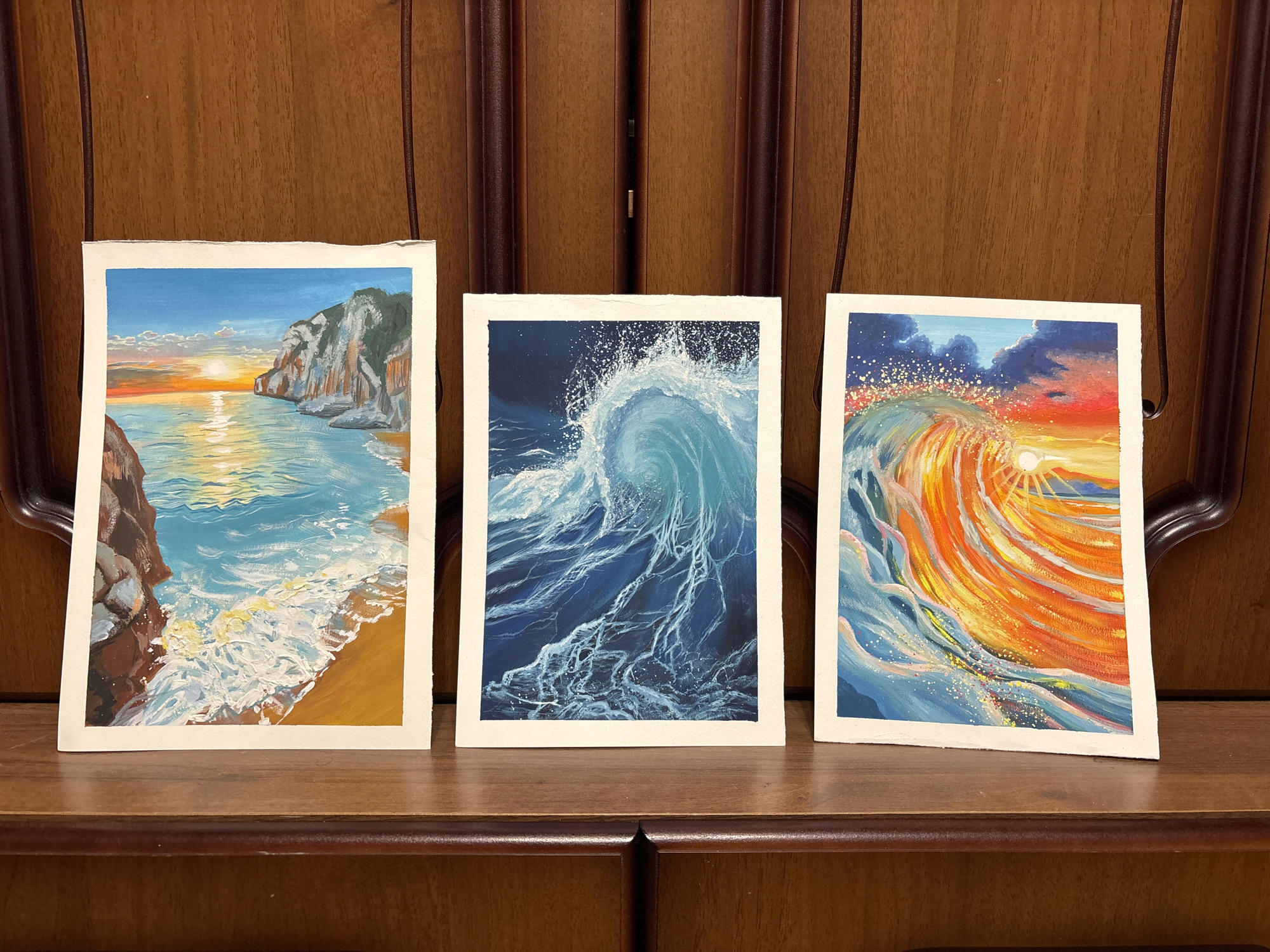 Three watercolor paintings are displayed on a dark brown wooden surface, likely a cabinet or shelf. The paintings are framed in white and feature coastal or ocean themes. From left to right:
1. A coastal landscape with a sunset. The scene includes cliffs, ocean water with reflections of the setting sun, and a small beach. The sky is a mix of blue, orange, and yellow hues.
2. A close-up of a breaking wave. The water is various shades of blue, with white foamy details. The background is a dark blue.
3. A stylized depiction of a wave with a sunset or sunrise in the background. The wave is rendered in shades of orange, yellow, and blue.
The setting appears to be indoors with the paintings arranged in a row against a wooden background. There is no indication of Izhevsk, Russia.
