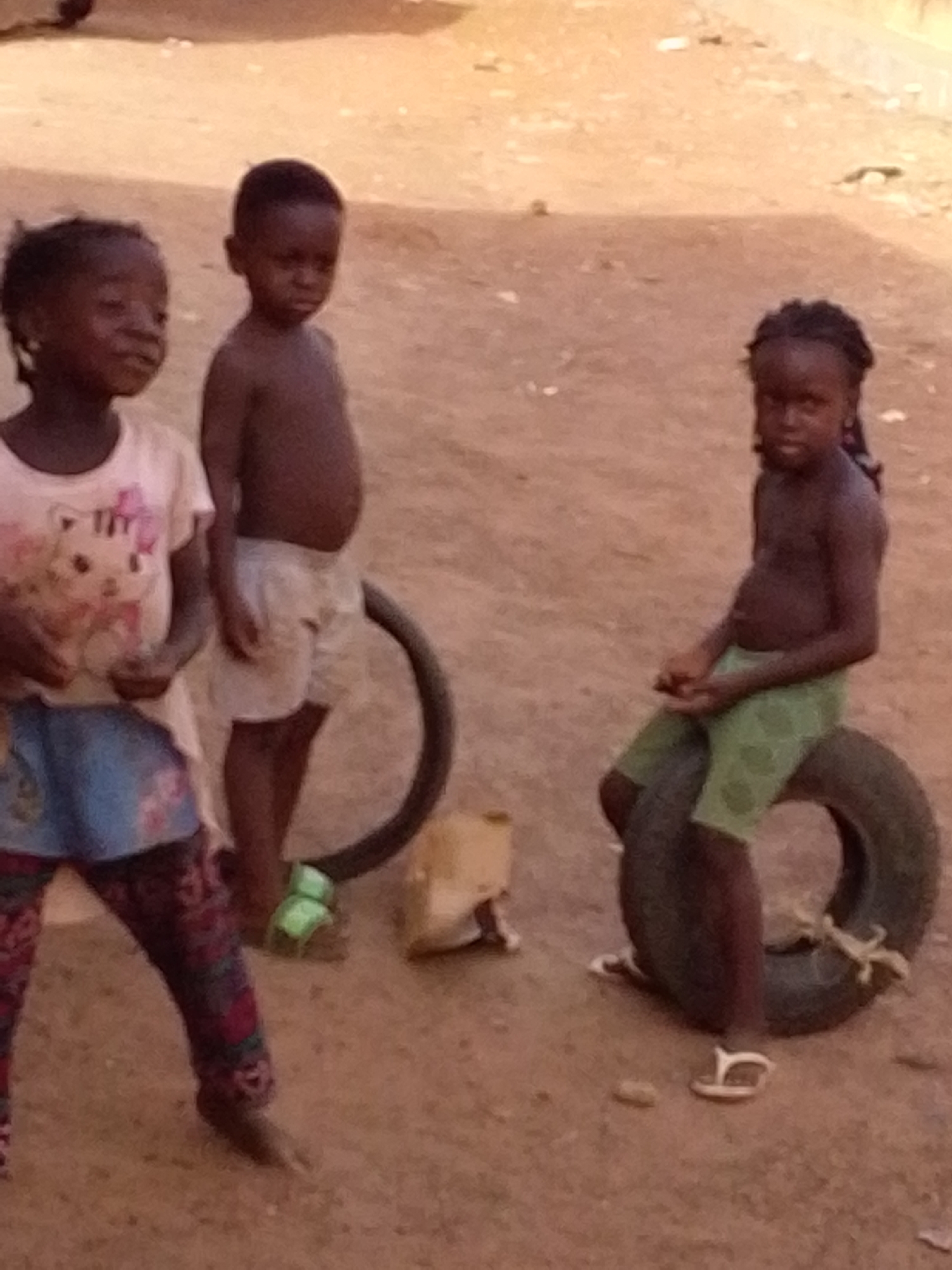 Three young children are present in the scene, two standing and one sitting on a tire. A girl to the left wears a light-colored t-shirt with a graphic and patterned pants. A boy stands next to her shirtless, wearing light-colored shorts. Another girl is sitting on a tire to the right, also shirtless, wearing green shorts. Another tire and some sandals are visible in the shot. The environment appears to be an outdoor area with a dirt or sand surface. The location is an unspecified city in an unknown country.