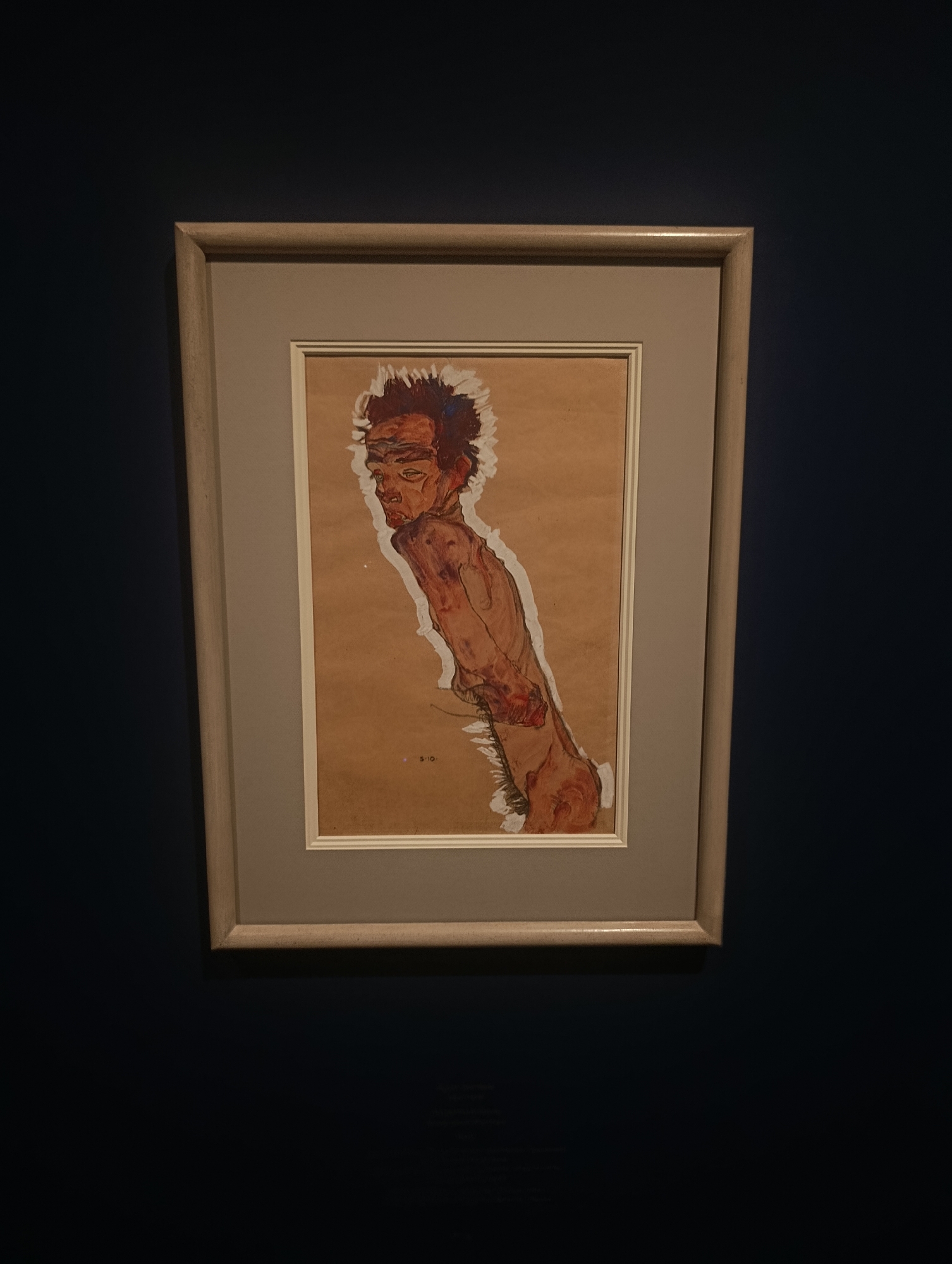 This media file depicts a framed drawing of a nude individual. The artwork is presented on a light brown paper, with the subject rendered in a style characterized by strong lines and expressive brushstrokes. The figure is shown from the side, with their head turned away and their body contorted. The artist has used a limited color palette, primarily earthy tones and reds, to depict the skin and musculature. The subject has dark, spiky hair and their facial features are sharply defined, conveying a sense of intensity or anguish. There is a stark white outline around the figure, separating it from the background and emphasizing its form.
The drawing is matted with a light gray border and encased in a simple wooden frame. The frame is light in color, likely natural wood, with a subtle grain. The entire piece is hung against a dark blue wall, creating a dramatic contrast that draws attention to the artwork.
Below the figure, in the lower right quadrant of the drawing, there appears to be a small, handwritten inscription that reads "S. 10.". Given the context of Vienna, Austria, and the style of the drawing, it is plausible that this is a work by Egon Schiele, a prominent Austrian Expressionist artist known for his raw and often unsettling portraits. The overall mood conveyed by the artwork is one of vulnerability and raw emotion. The lighting in the photograph suggests an indoor setting, possibly a gallery or a museum. There are no indications of specific time of day or weather conditions.