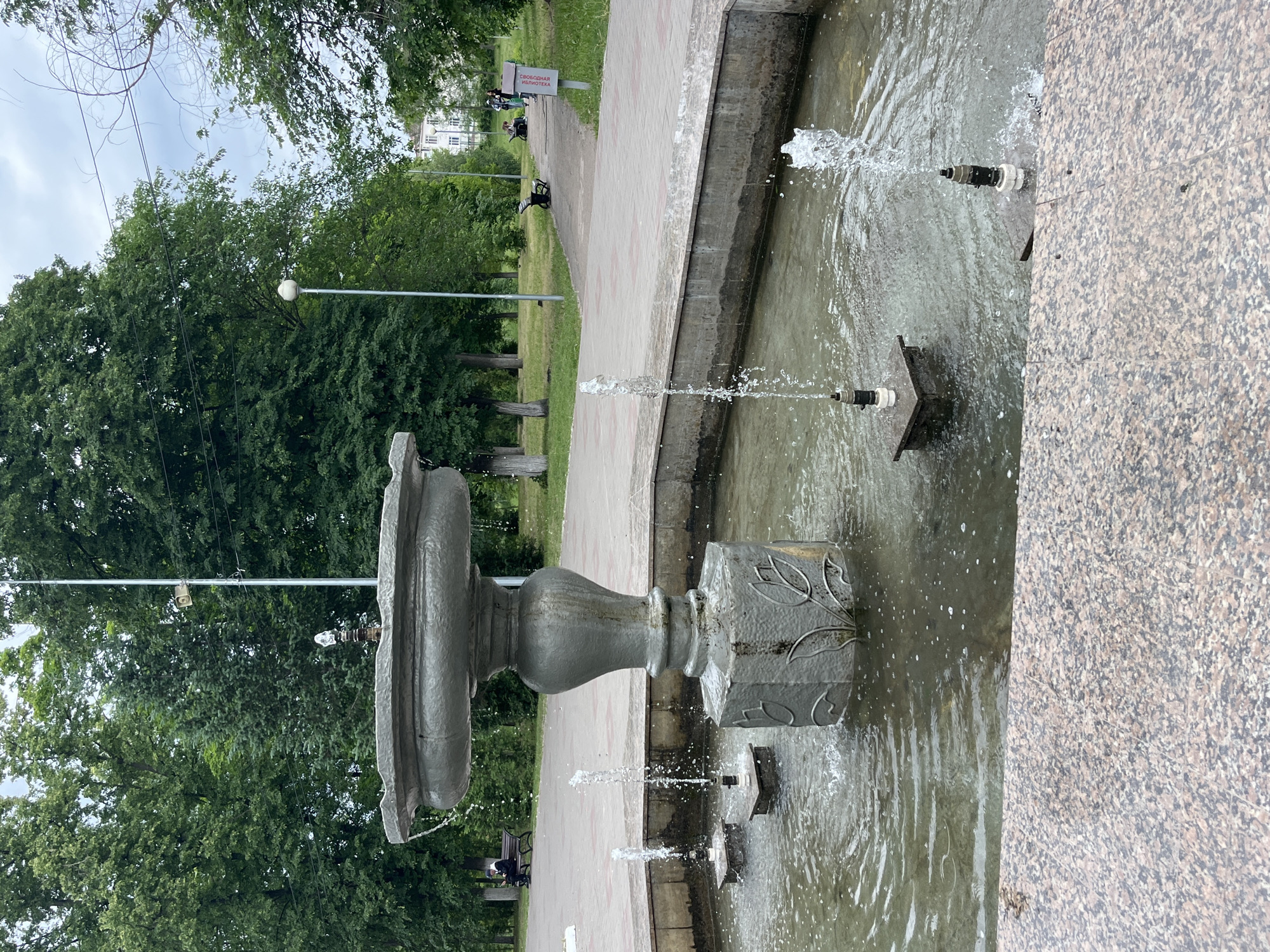 This media file captures a view of a public park or square, featuring an active fountain and lush greenery. The image is vertically oriented (when viewed without rotation).
In the foreground, a multi-tiered fountain is prominently displayed, actively spraying water from several nozzles. The main structure of the fountain is made of gray stone or concrete, with decorative carvings, possibly leaf patterns, on its base. Water splashes from the fountain into a shallow basin, which is part of a larger concrete structure. To the right of the fountain, a granular, light brown and gray tiled surface, likely part of a walkway or plaza, is visible.
Beyond the fountain, a light-colored, possibly cobblestone, path curves through the scene. On the left side of the path, several dark green deciduous trees create a dense canopy. Along the path, two dark benches are visible, with a few people sitting on them in the distance. One person is distinctly visible sitting on a bench to the left of the path.
A white sign with red lettering is positioned near one of the benches, displaying the Russian text "СВОБОДНАЯ БИБЛИОТЕКА," which translates to "FREE LIBRARY." This sign indicates a community initiative or amenity in the park.
In the background, behind the trees and path, parts of light-colored buildings with multiple windows can be seen, suggesting an urban or suburban setting. The sky is overcast or cloudy, indicating a daytime setting, possibly a mild or humid day given the green foliage. Several power lines or wires are visible crisscrossing the upper part of the sky. A streetlight with a spherical lamp is also visible among the trees.
The scene depicts a peaceful public space where people can relax and enjoy the environment, with the fountain providing a focal point and the "Free Library" sign adding a community-oriented detail.
