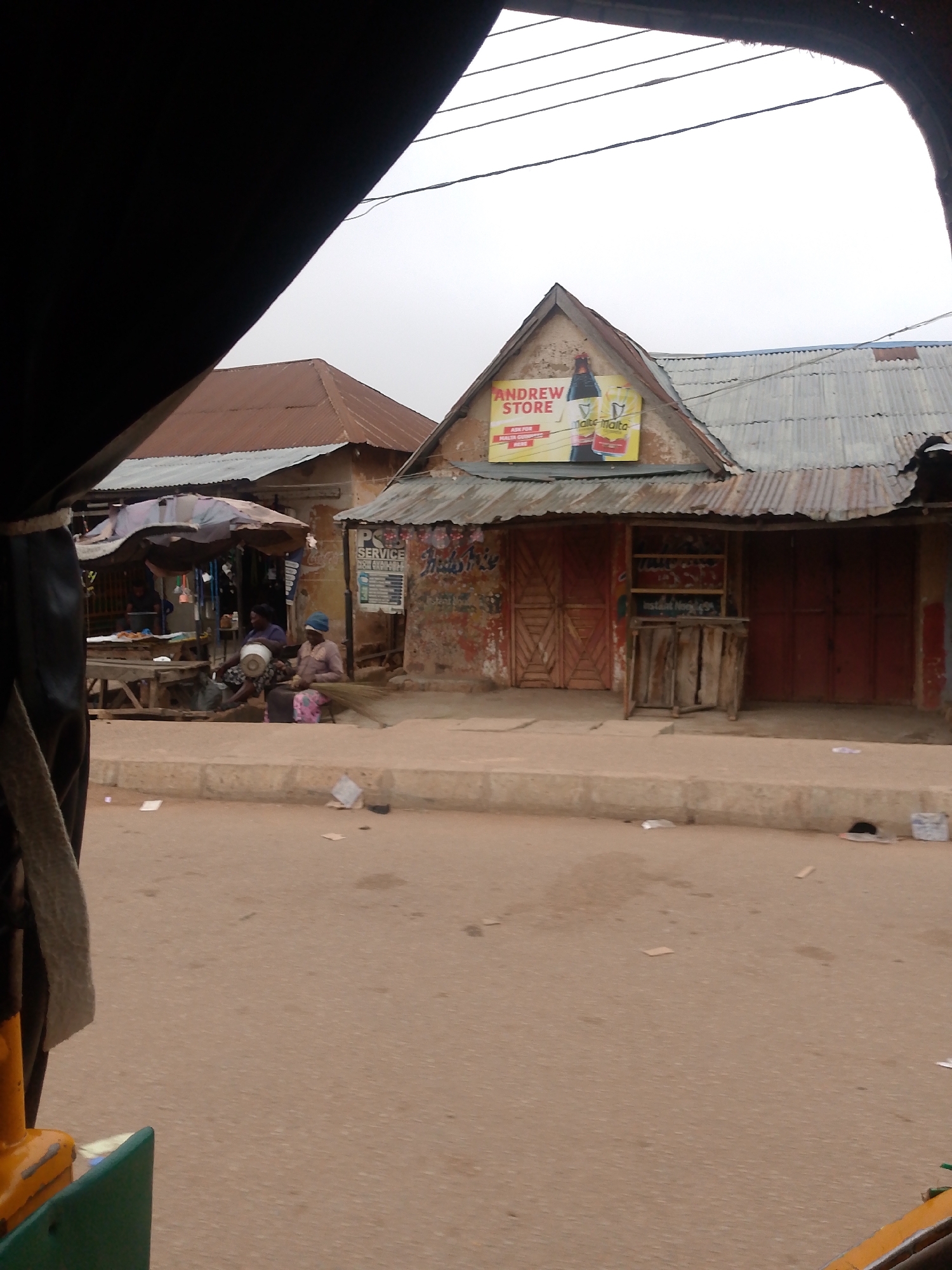 The scene is set in Bukuru, Nigeria, showing a street view from a vehicle. A building with a peaked roof features an "Andrew Store" sign advertising Guinness and Malta drinks. The building has weathered, corrugated metal roofing, red doors, and a small display case. Adjacent to it, another structure with a similar roof and signage is visible.
Two women are sitting on the sidewalk near a market stall under an umbrella, engaging in an activity involving a metal container and other objects. The street in the foreground is paved and littered with debris. A portion of the vehicle from which the photo was taken is visible in the lower left and upper left corners of the image. Electrical wires stretch across the upper part of the scene against a cloudy sky.
